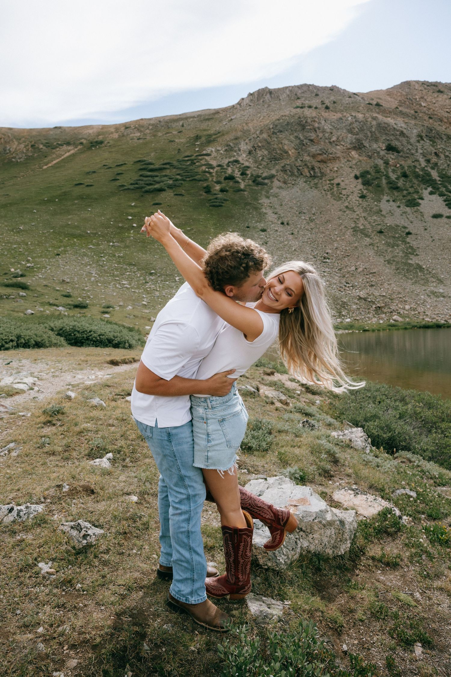 Couple embracing and laughing near a mountain lake, woman in denim skirt and cowboy boots, green hills backdrop.