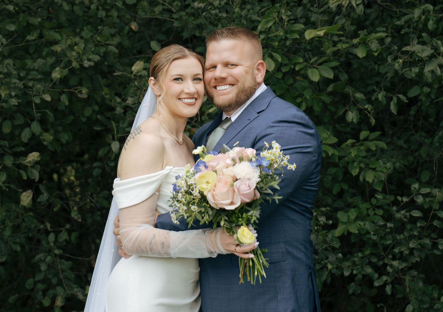 Smiling bride and groom pose together on their wedding day, she holds a pastel bouquet, he wears a navy suit.