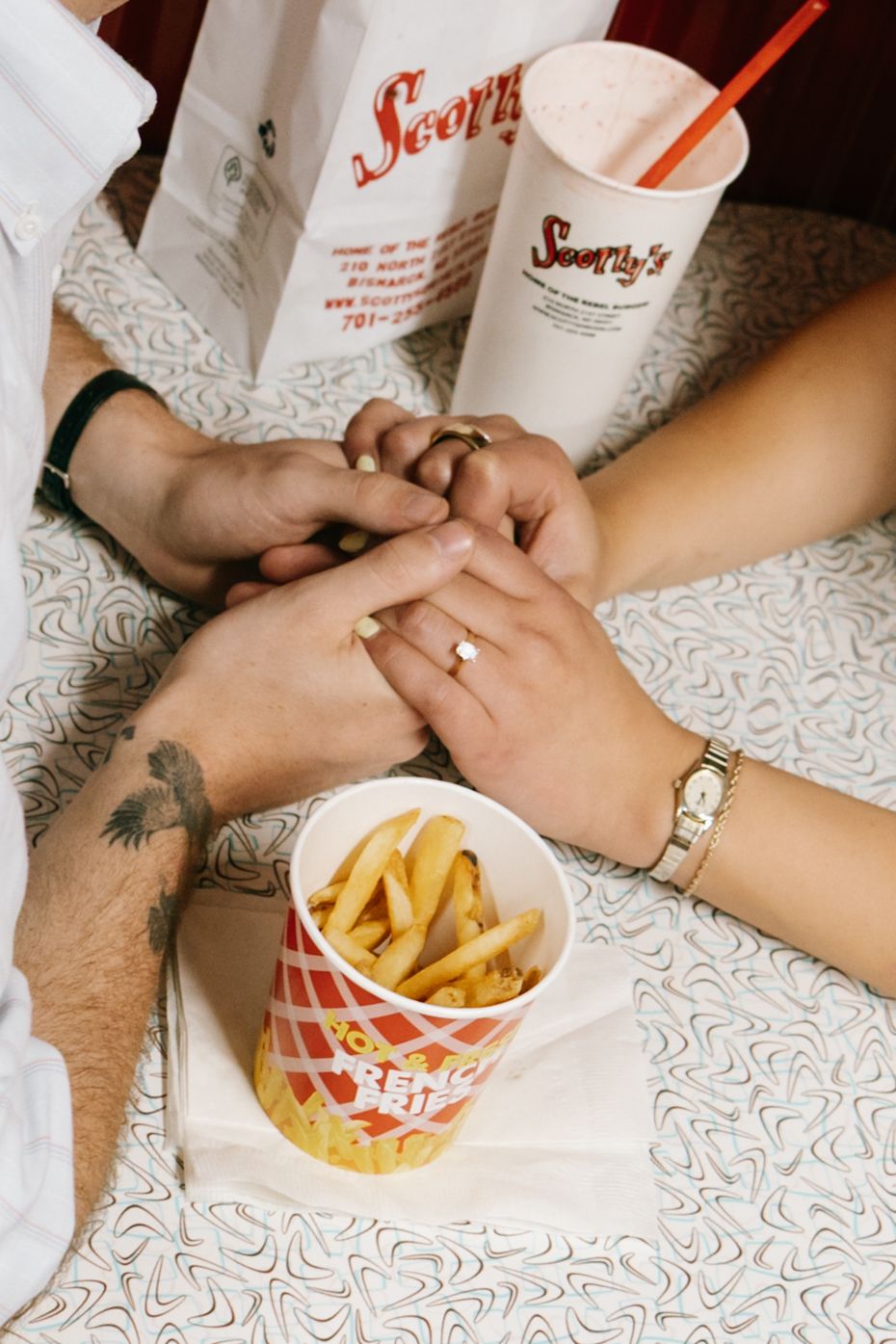 Couple holding hands with engagement ring at Scooter's, sharing fries and a milkshake on a white lace table.