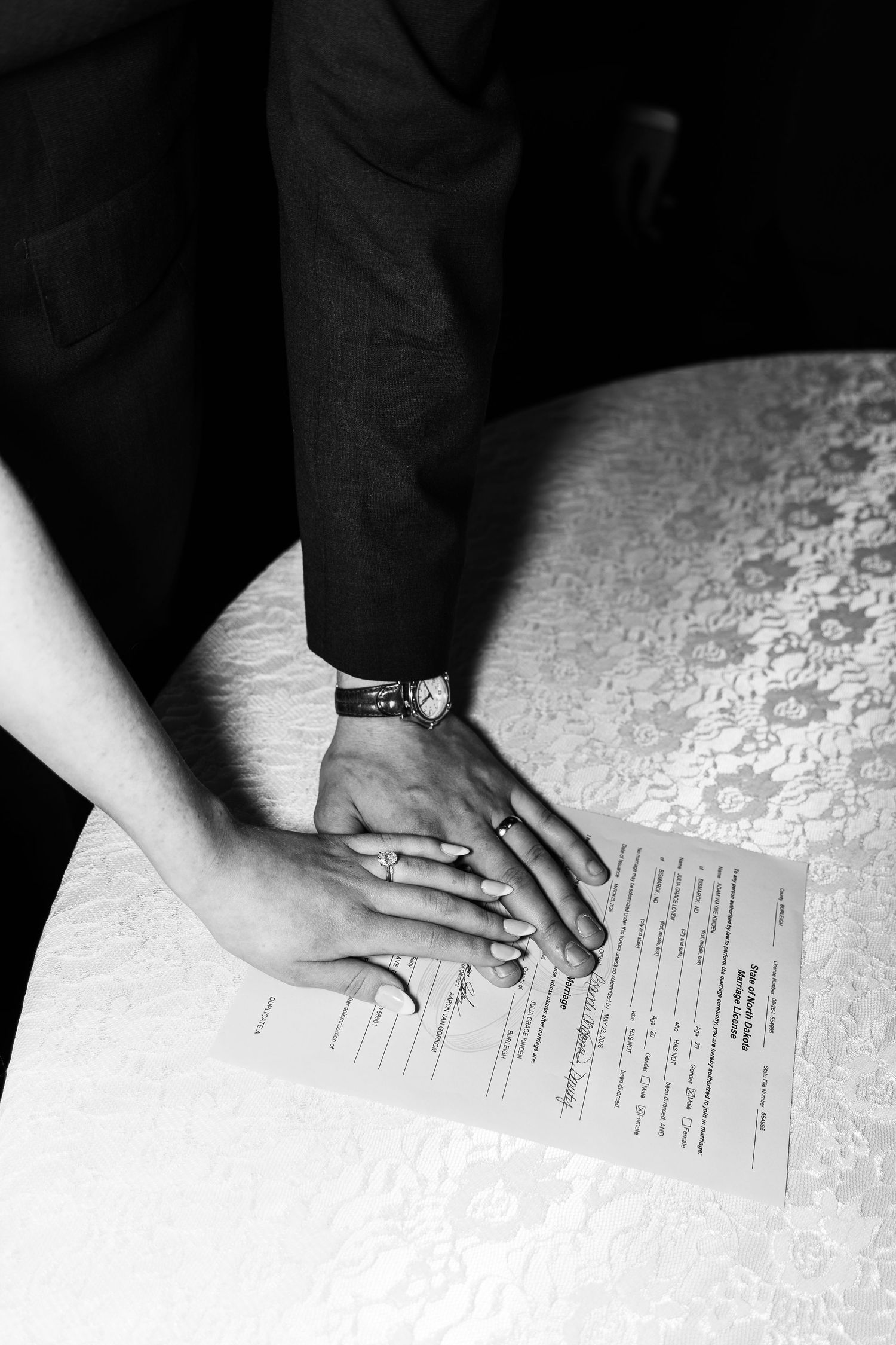 Black and white photo of newlyweds placing hands on marriage certificate, showing wedding rings and watch.