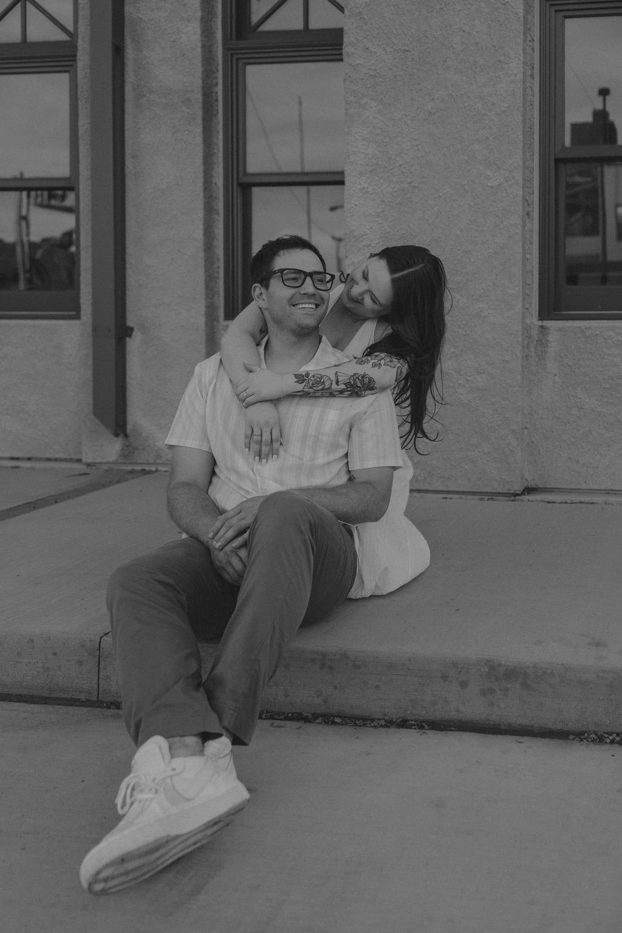 A romantic black and white portrait of a couple sharing a tender moment while sitting on steps outside a building.