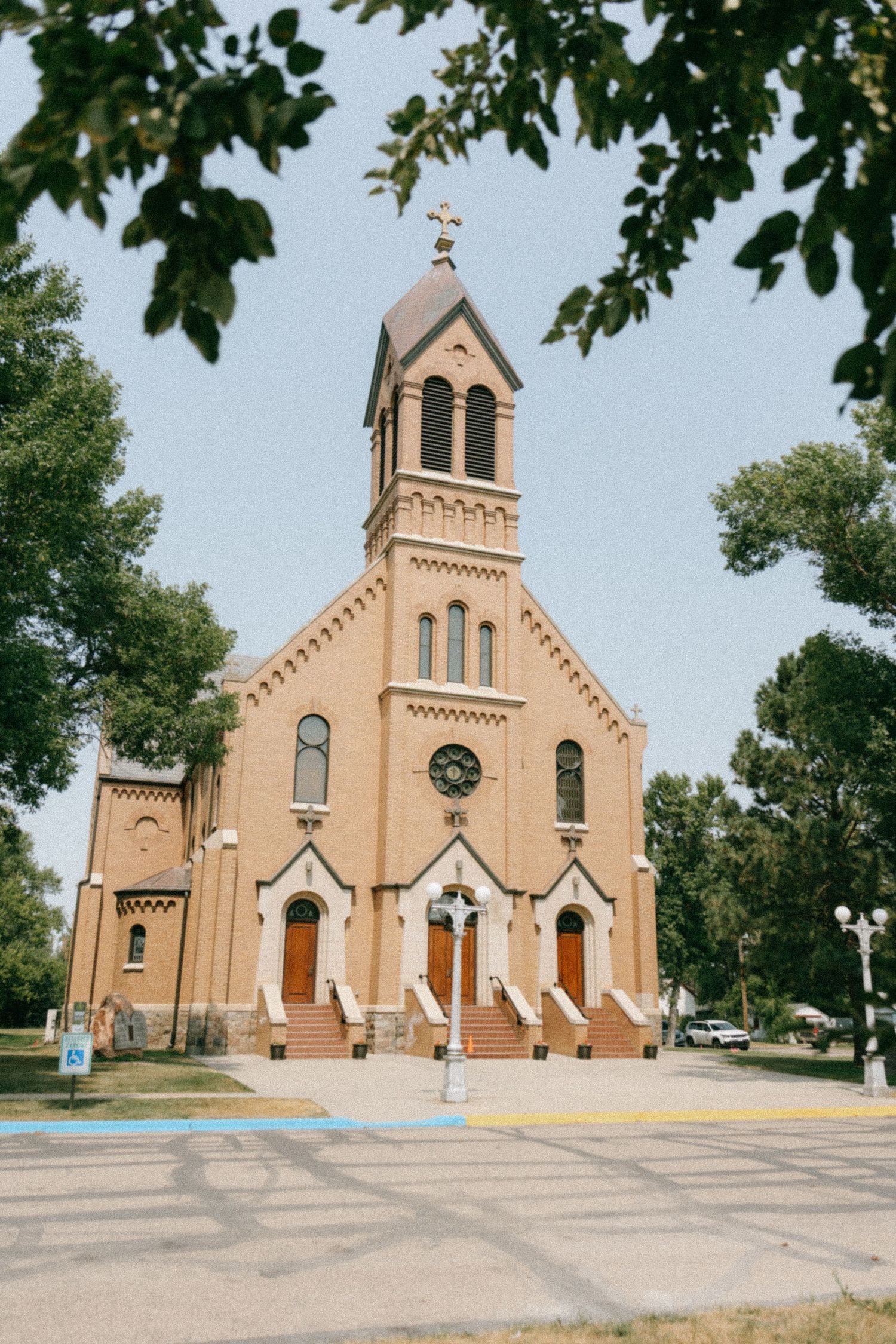 Historic tan brick Catholic church with bell tower and cross, framed by green trees on a sunny day.