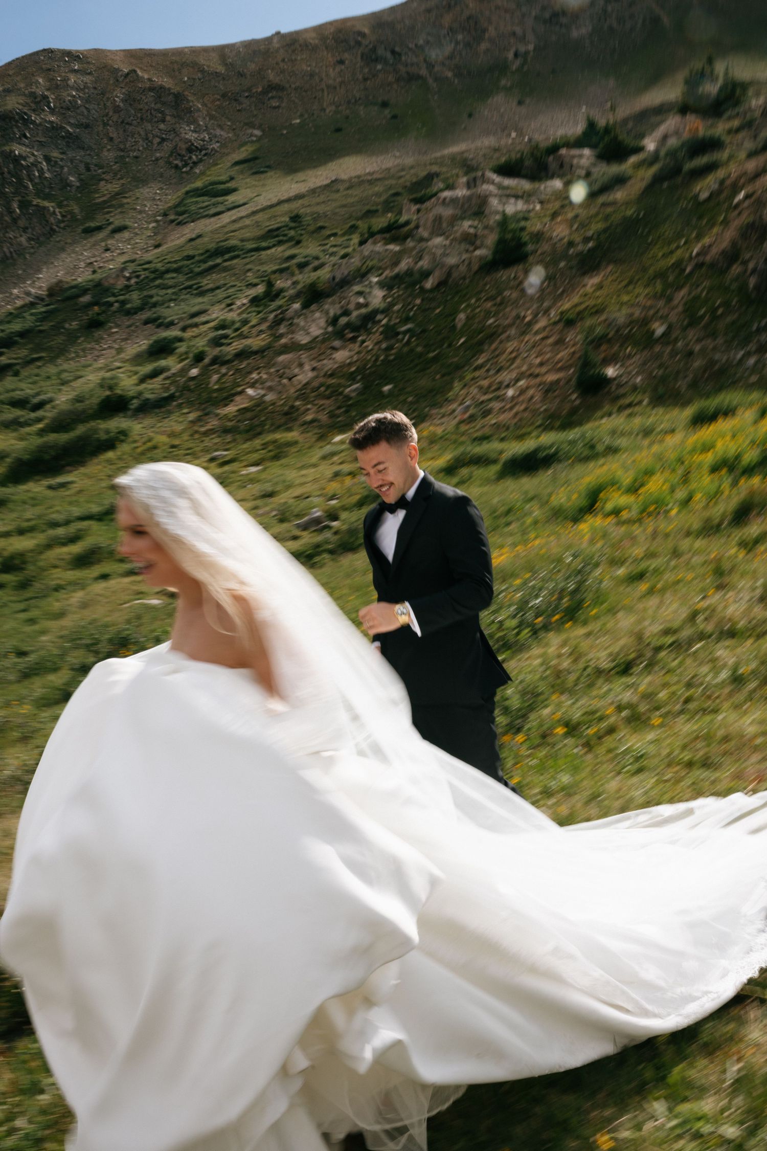 Bride in flowing white gown and groom in black tuxedo walking through green mountain meadow on their wedding day.