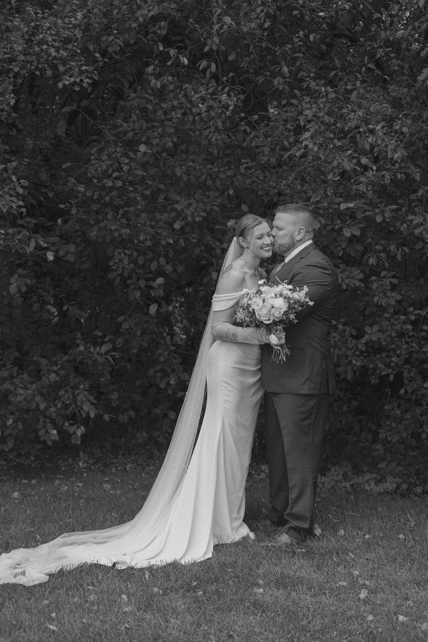Black and white wedding photo of couple embracing outdoors with long dress train.