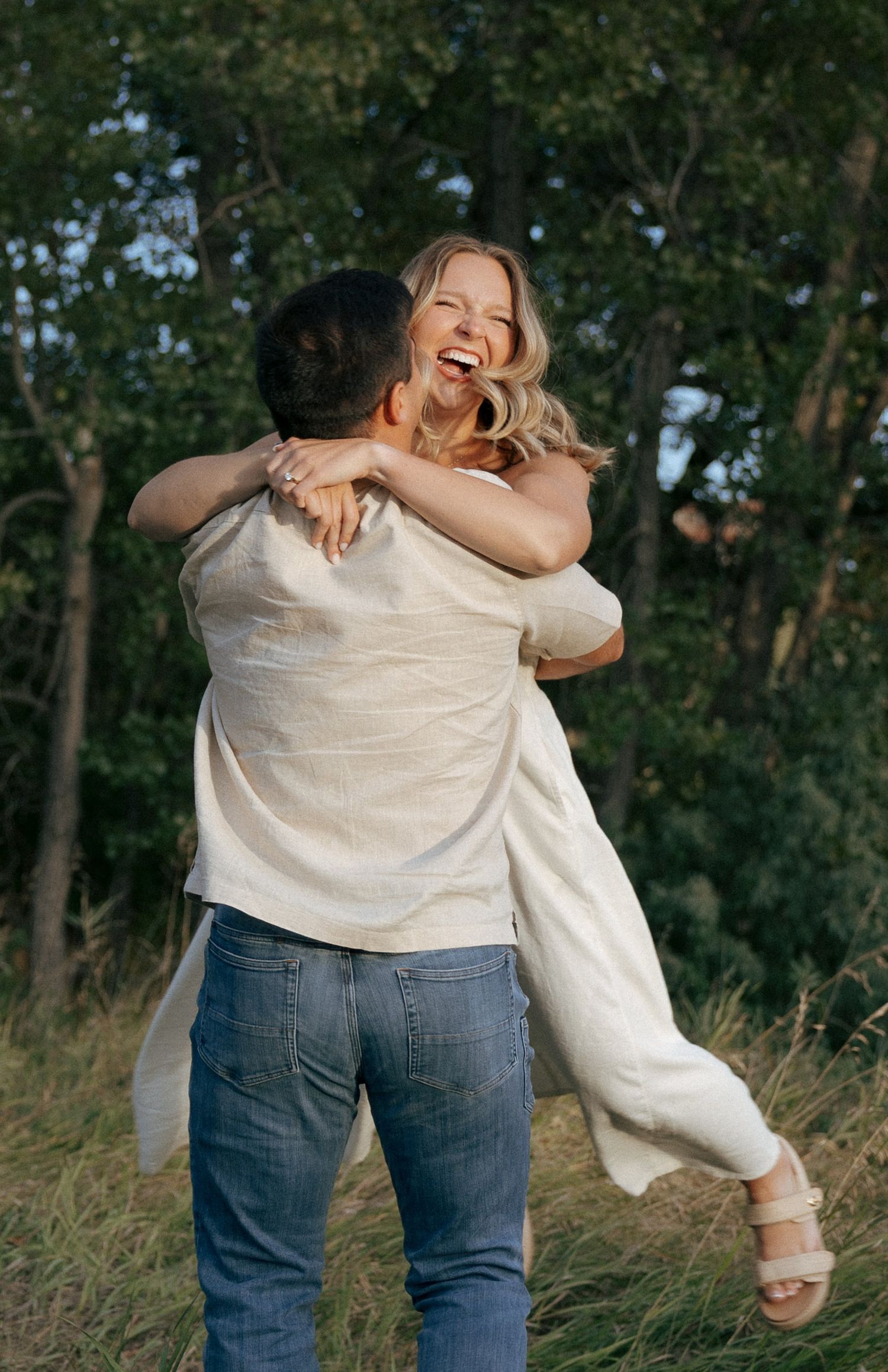 Two people share a playful embrace while dancing in tall grass near a wooded area.