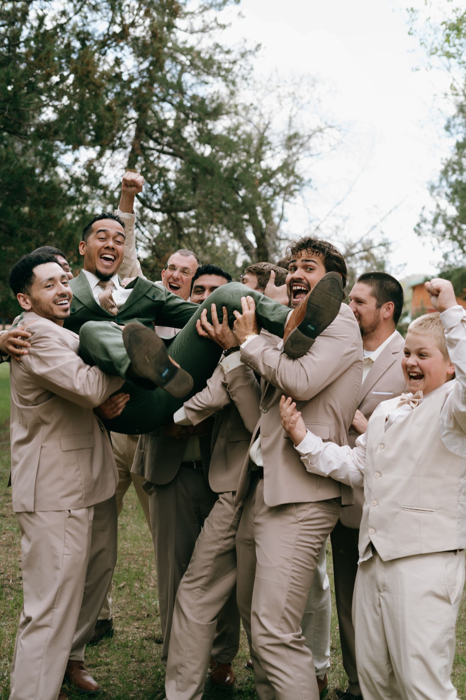 Groomsmen in tan suits joyfully lift the groom in a green suit during a fun outdoor wedding celebration.
