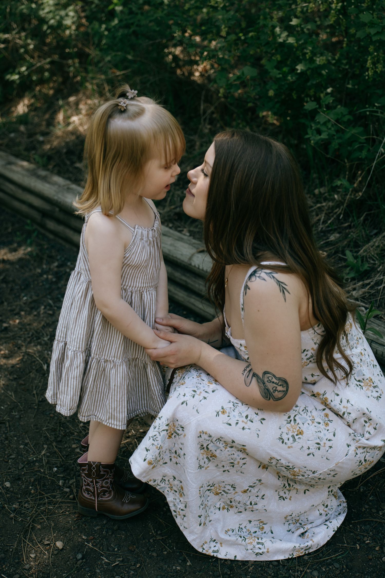A mother and toddler daughter share a tender nose-to-nose moment outdoors, both wearing summer dresses.