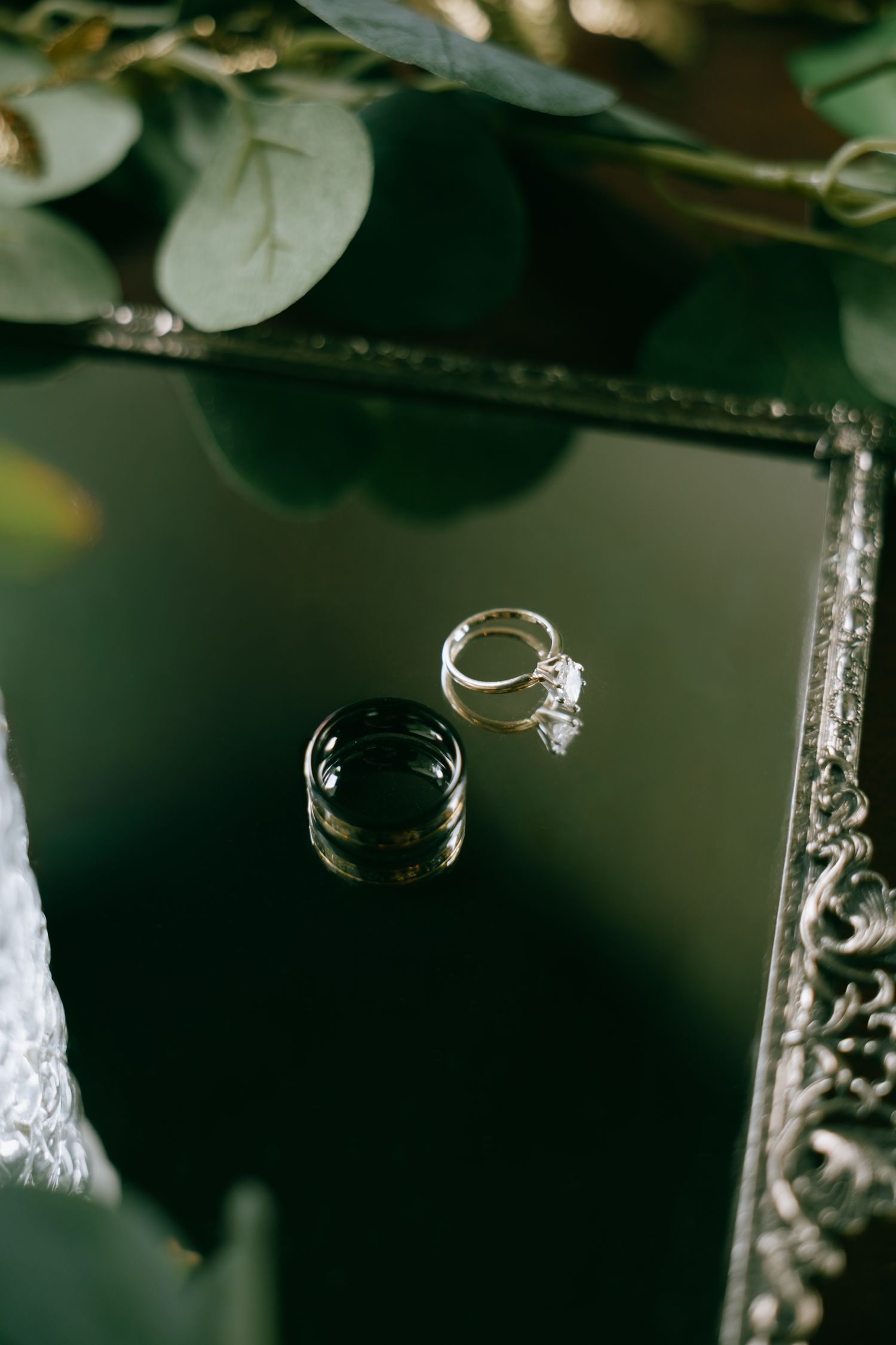 Diamond engagement ring reflected on a dark green mirror surface surrounded by eucalyptus greenery.