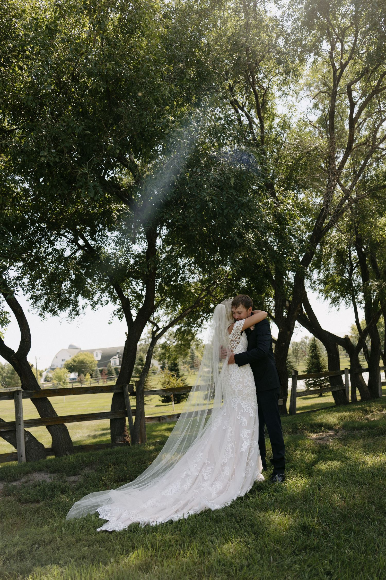 Bride and groom share a romantic kiss outdoors, with her lace gown and veil flowing in a lush green garden.