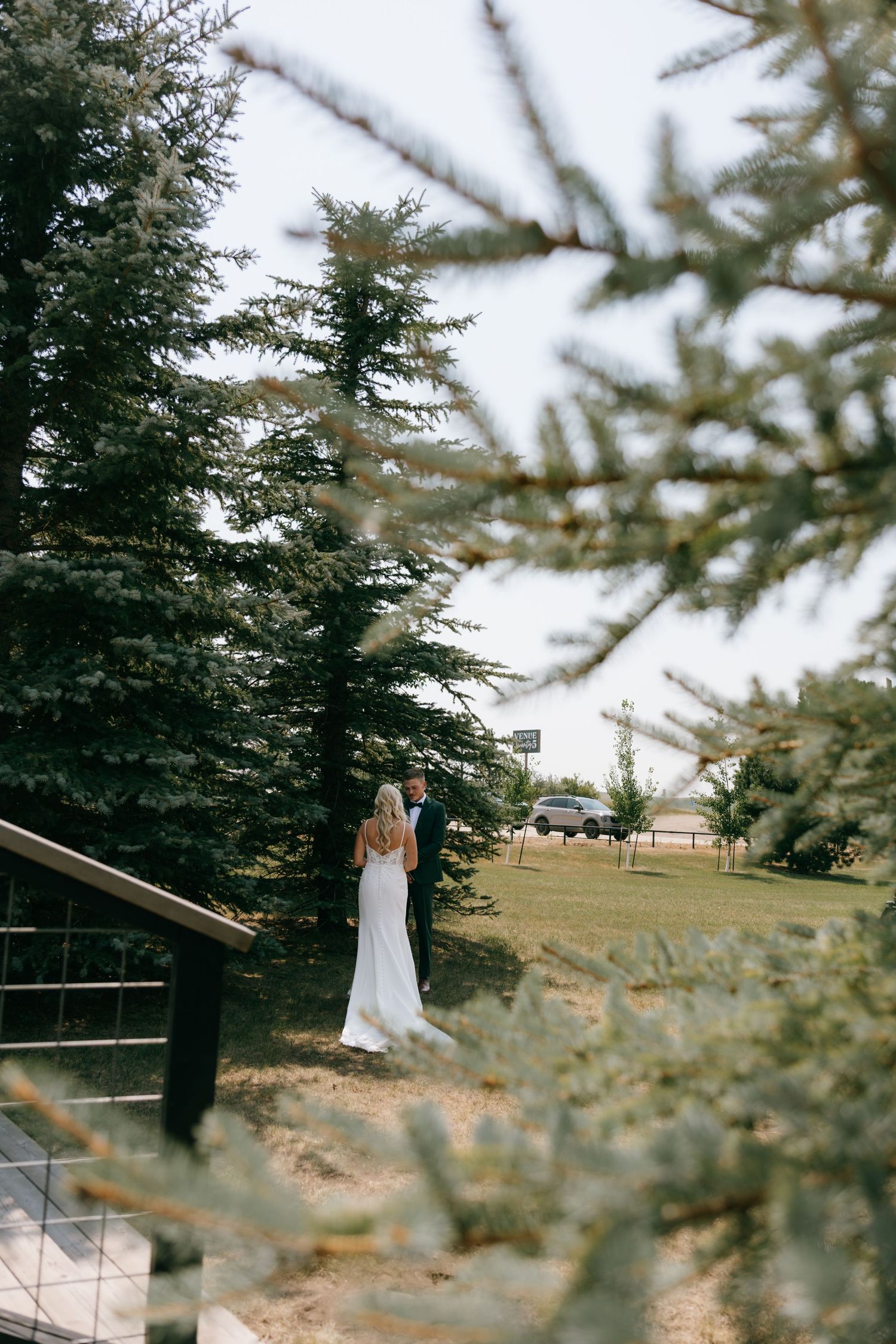Bride and groom standing among tall evergreen trees at an outdoor wedding venue on a cloudy day.