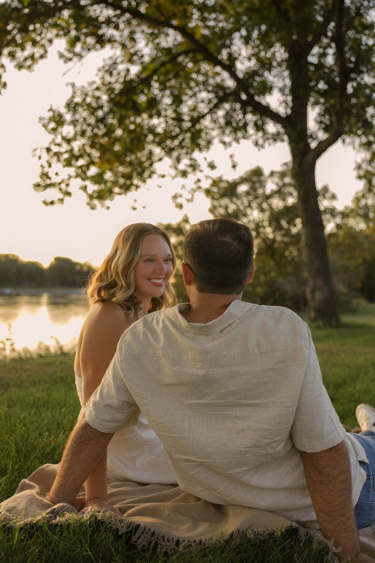 A couple shares a tender moment while sitting on grass near a lake at sunset.