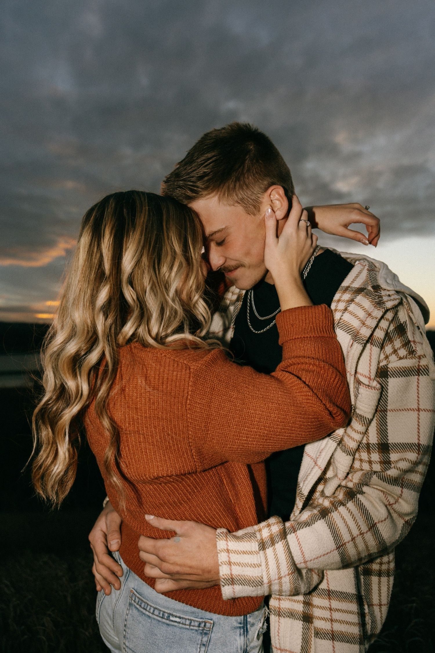 Tender moment between couple in rustic attire against sunset sky.