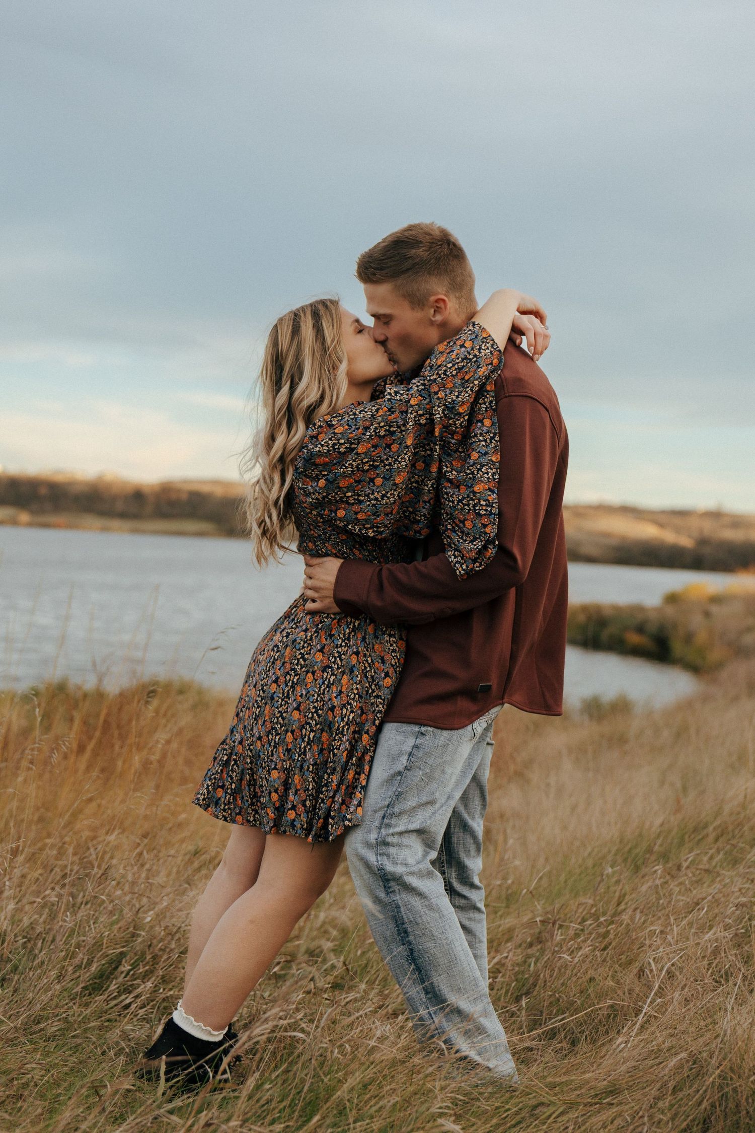Couple in casual wear sharing intimate moment on grassy hillside by water.