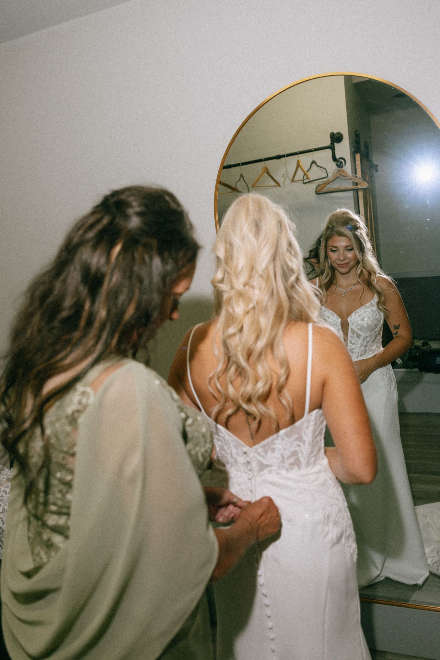 Bride in white lace gown views her reflection in a gold mirror while bridesmaid assists with dress.