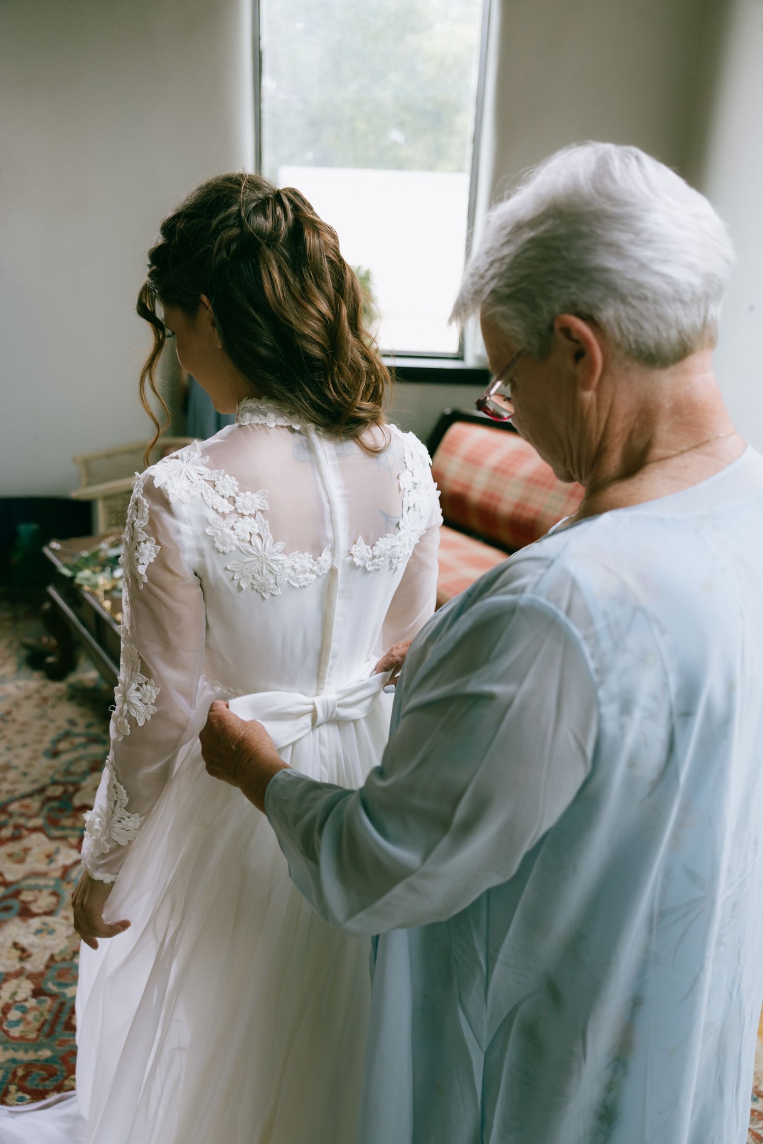 An older woman helps a bride fasten her lace wedding gown from behind near a bright window.