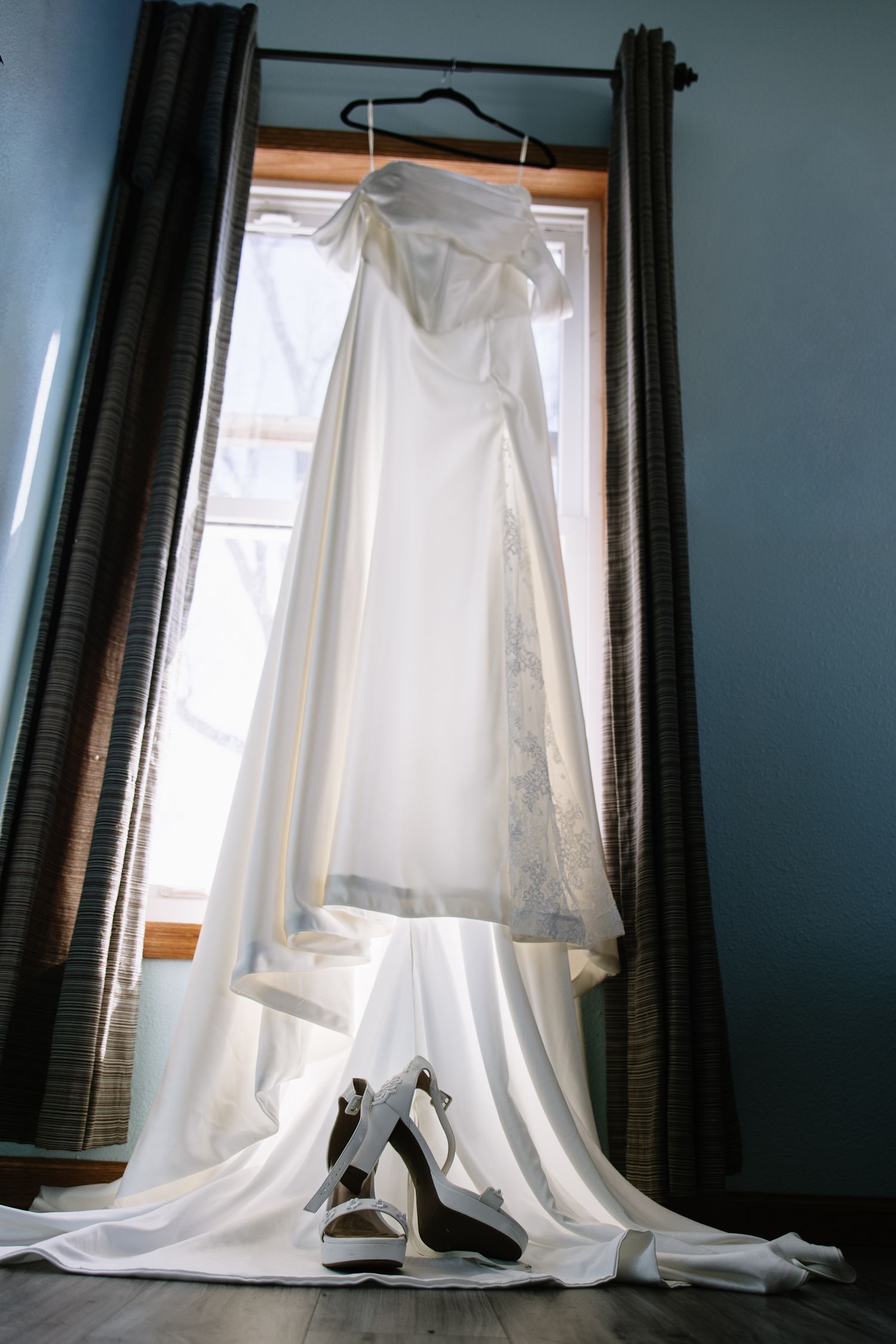 White wedding dress hanging in window with bridal heels below, elegant bridal preparation scene.