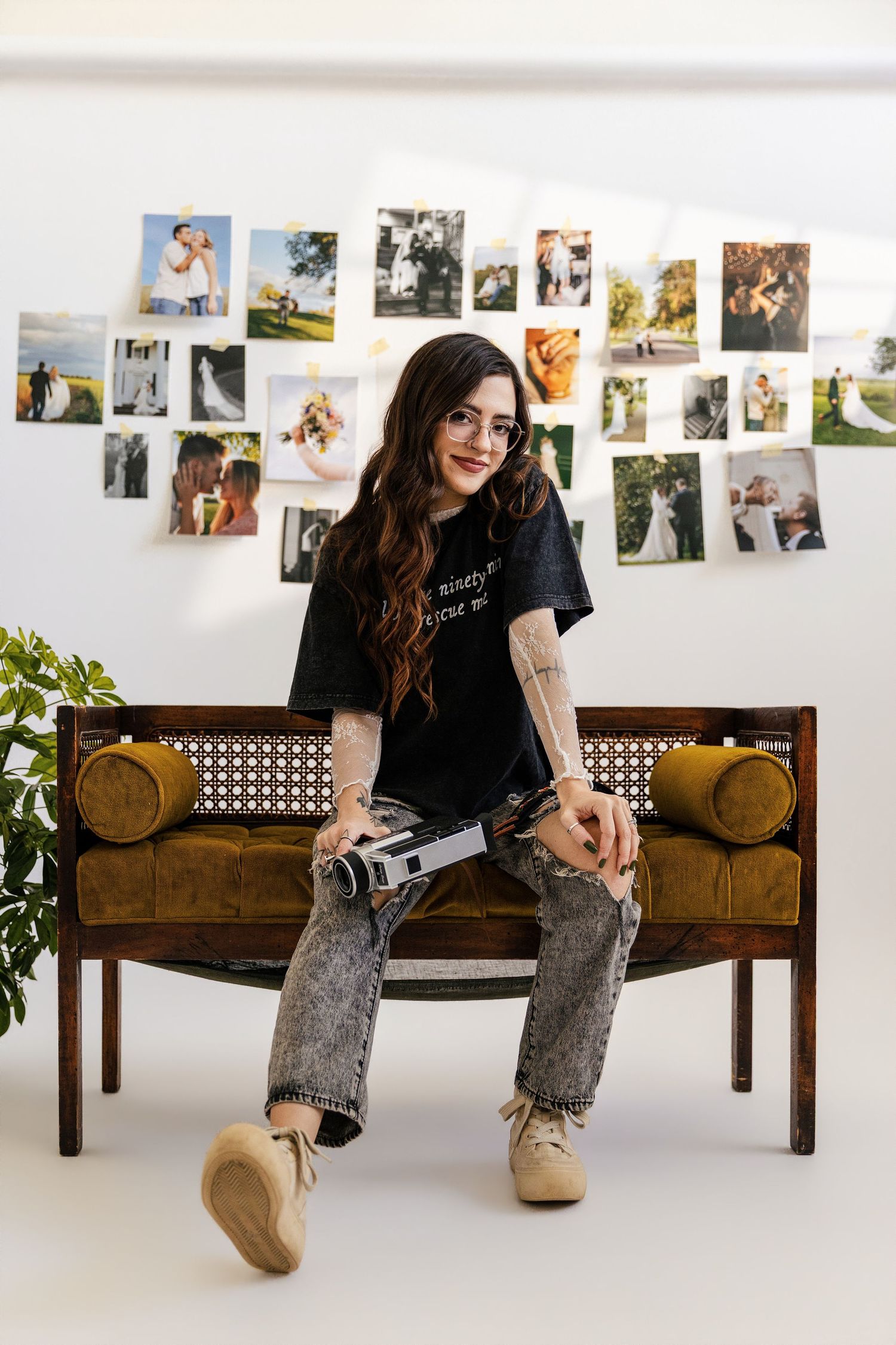A tattooed woman with long hair sits on a vintage bench holding a camera, with wedding photos displayed on the wall behind her.