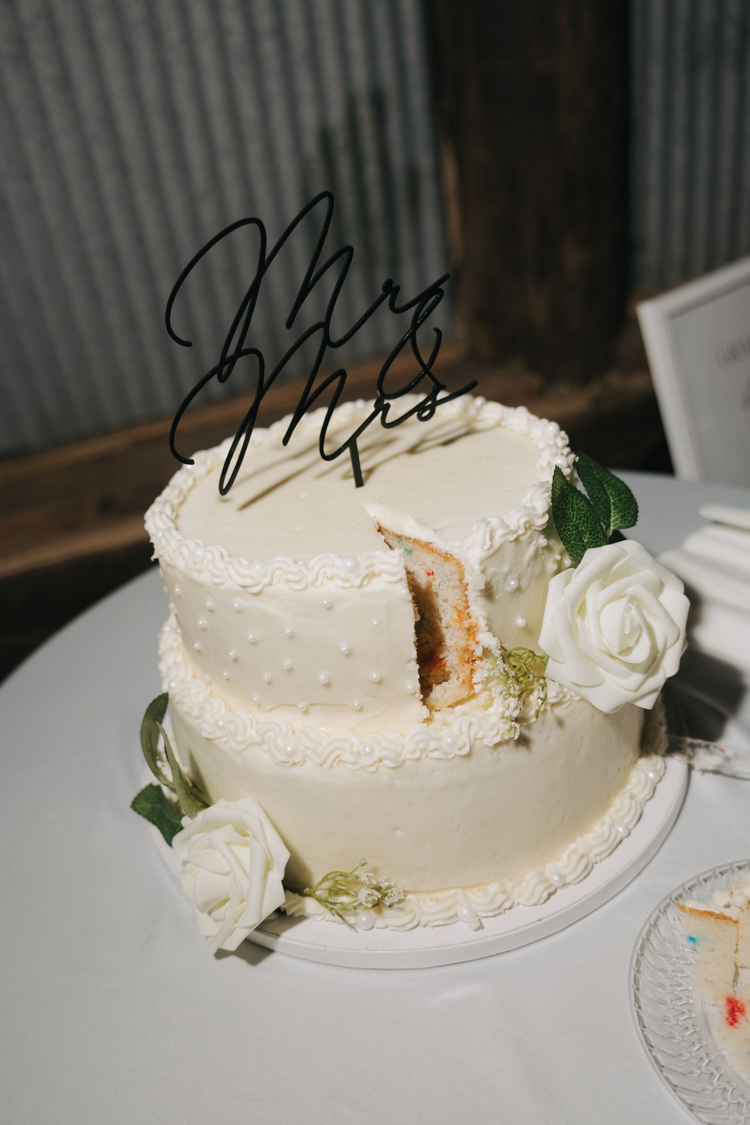 Two-tier white wedding cake with Mr & Mrs topper, white roses, and a slice cut out revealing golden layers inside.