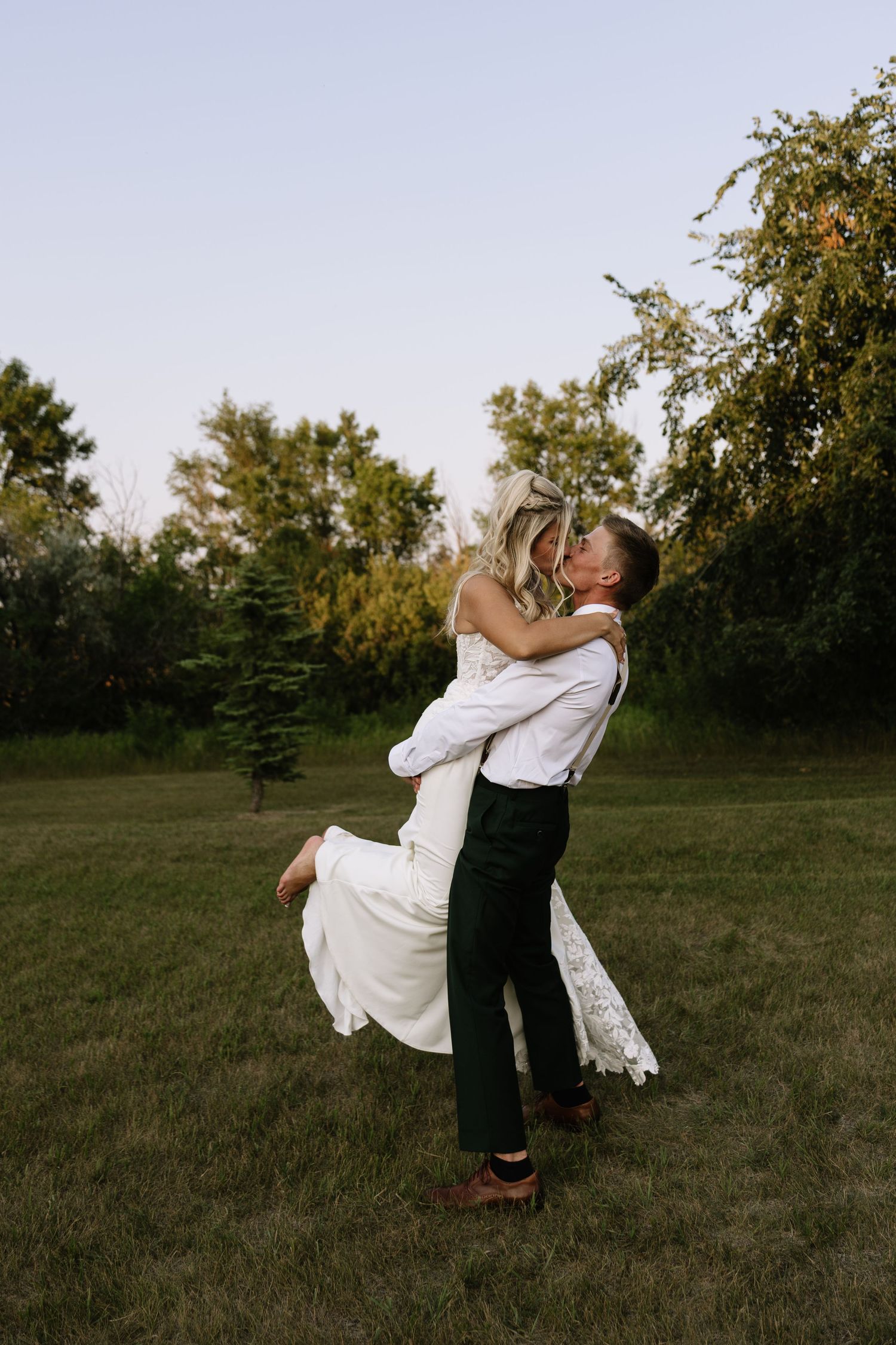 Groom lifts bride in white lace dress for a romantic kiss outdoors on green grass with trees in background.