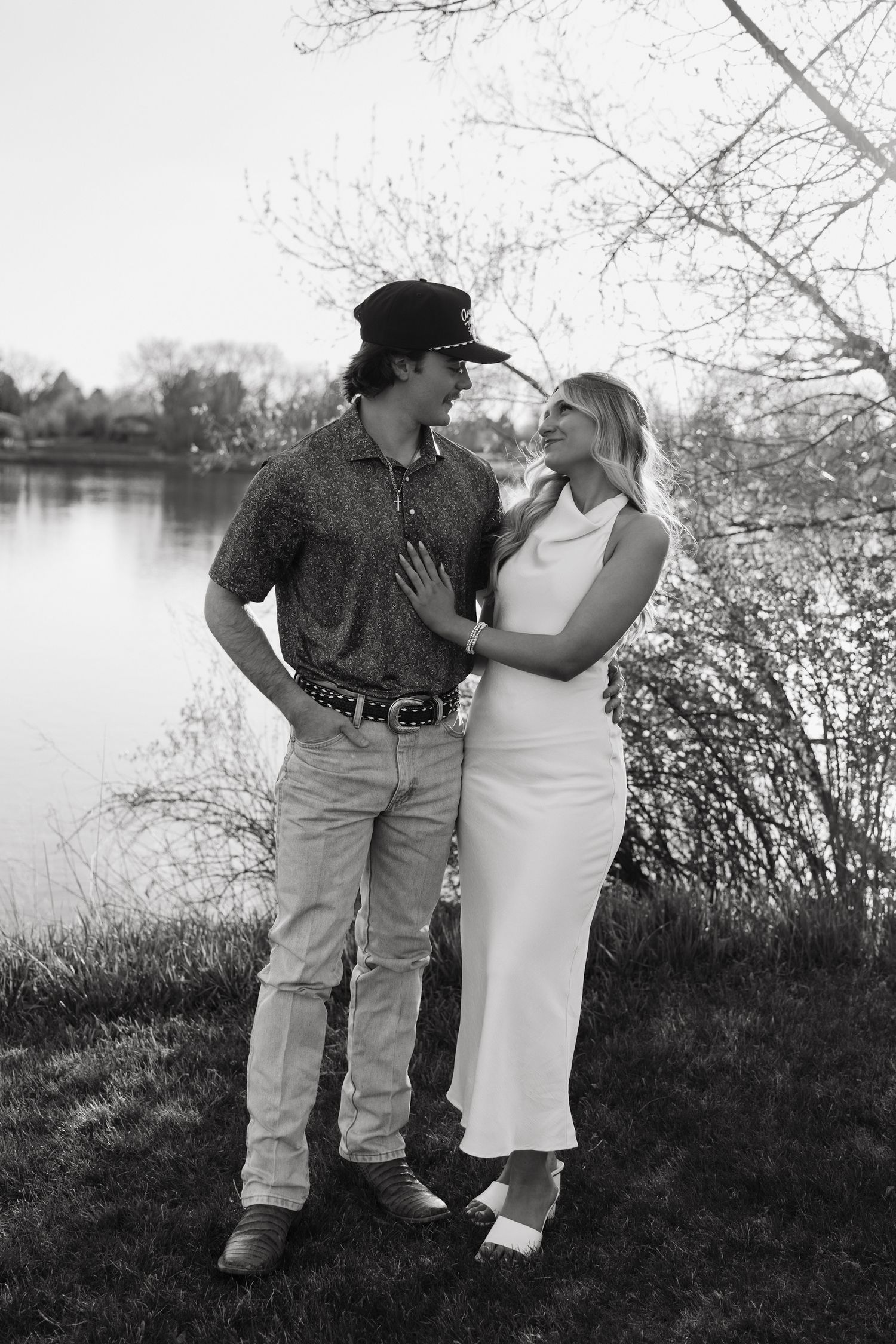 Black and white photo of a couple gazing at each other near a lake with blooming trees.