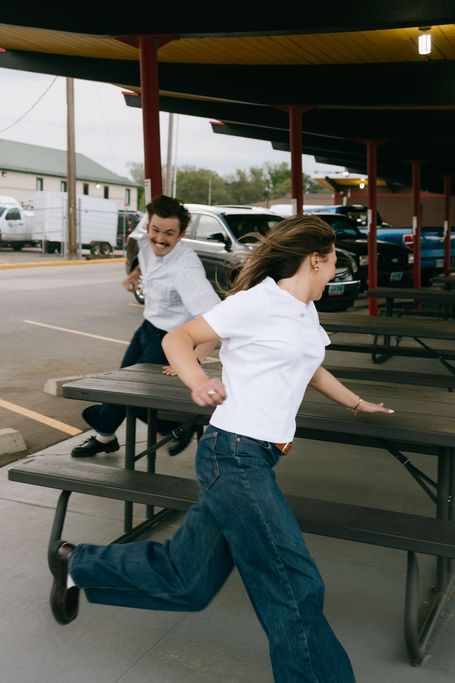 A man and woman in white shirts and jeans playfully chase each other around picnic tables at an outdoor restaurant.