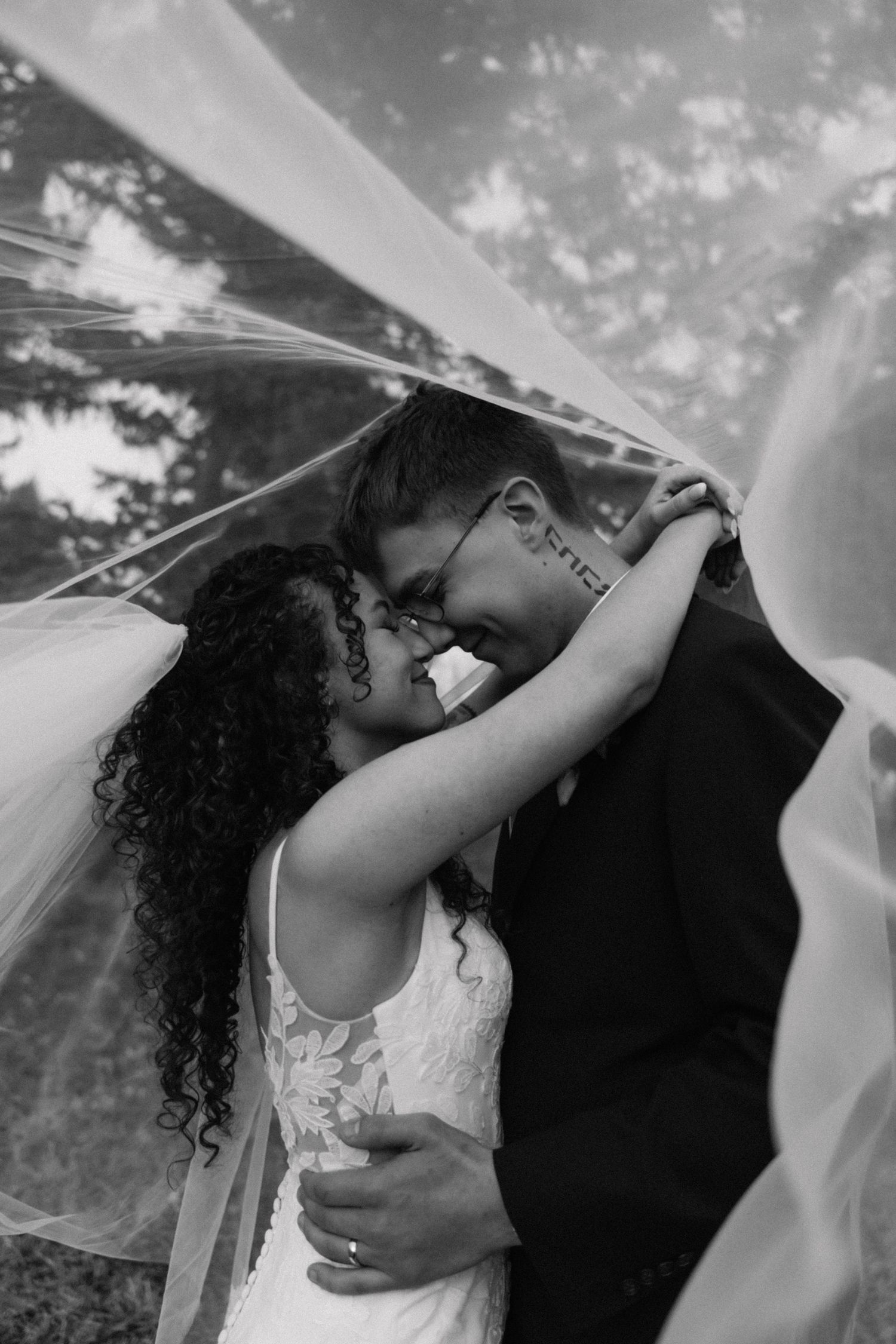 Black and white photo of a bride and groom sharing a romantic moment under a flowing veil outdoors.