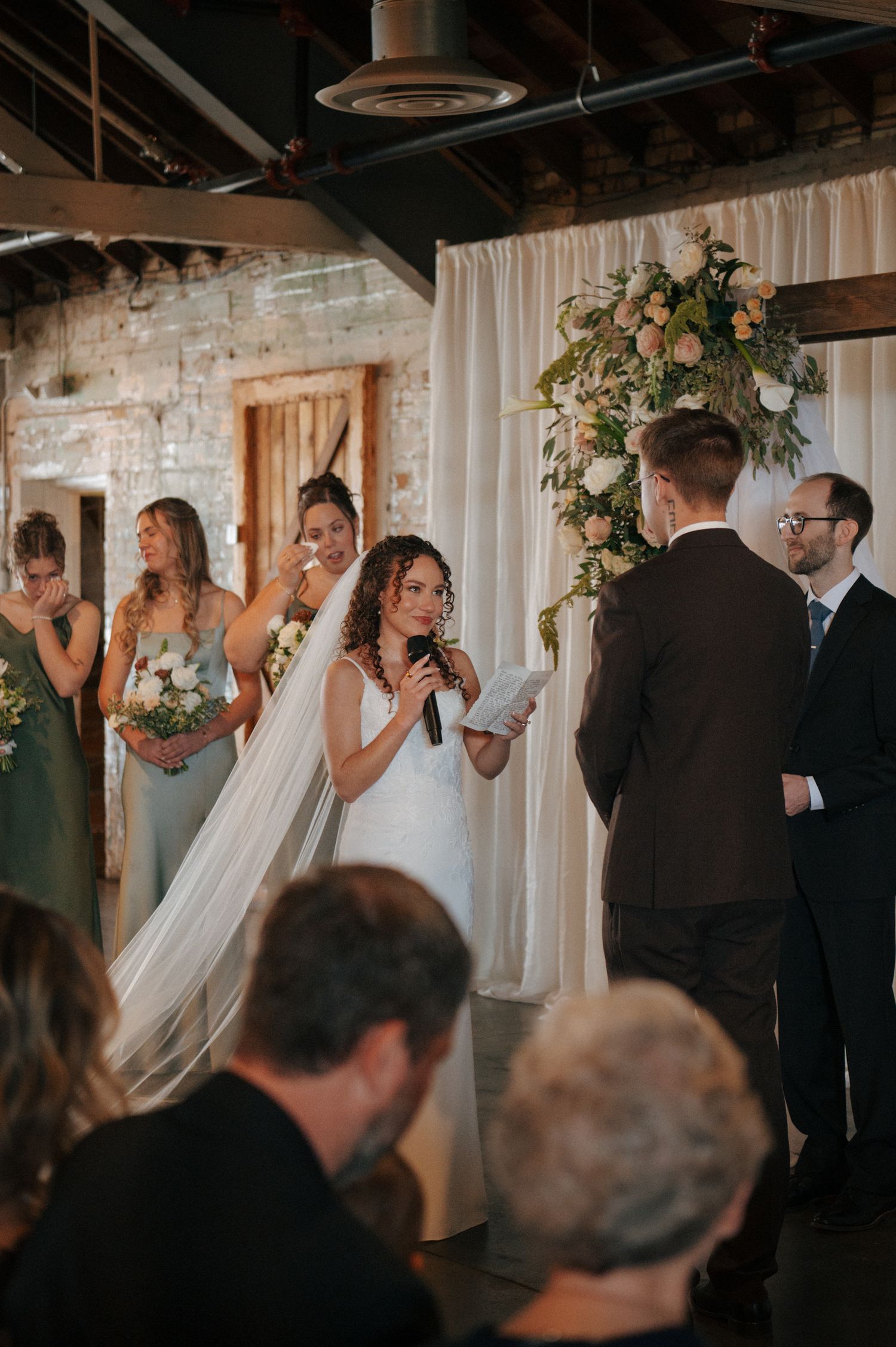 Bride reads vows at indoor wedding ceremony with bridesmaids in sage green dresses and floral arch backdrop.