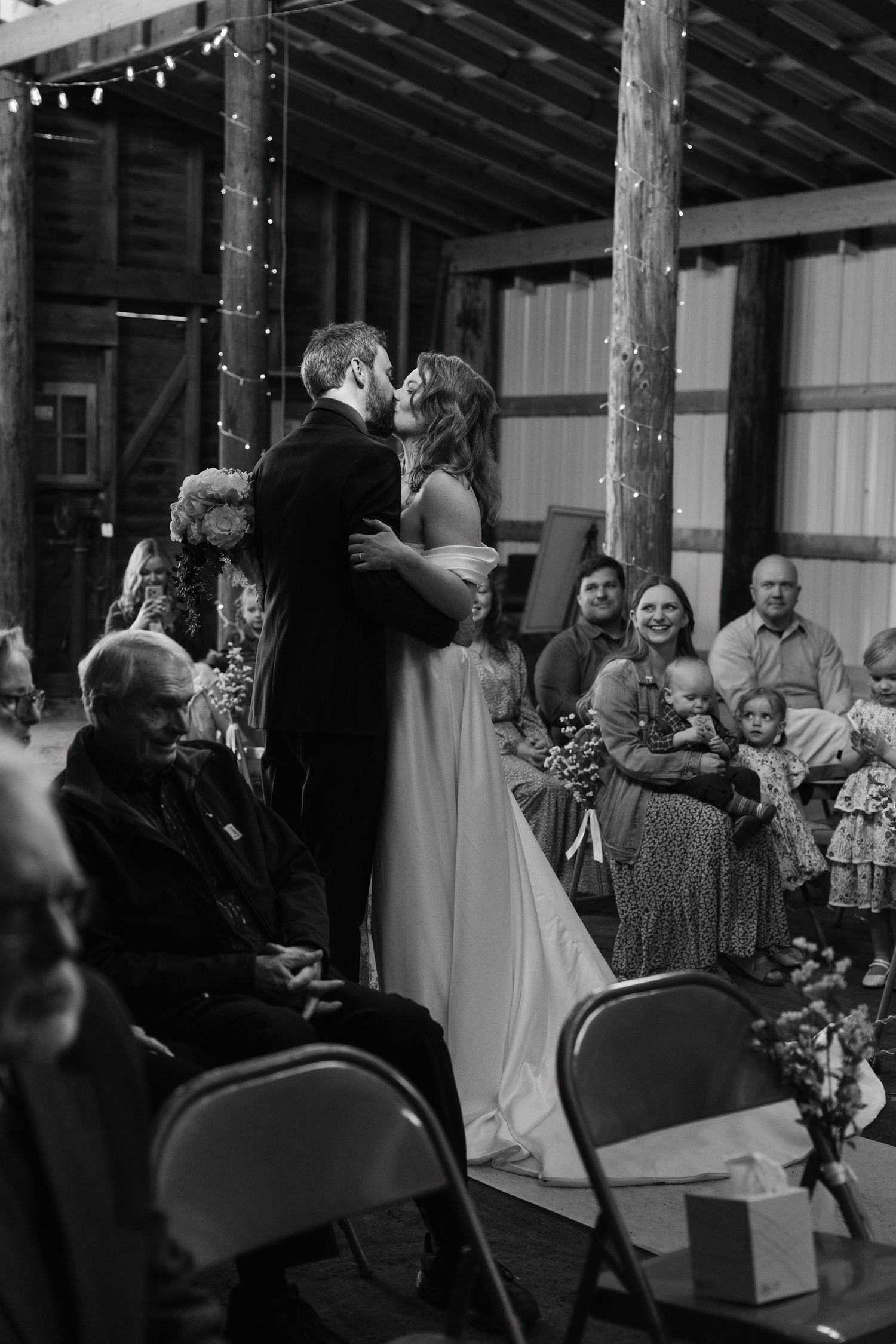 Black and white photo of a couple sharing their first kiss at a rustic barn wedding ceremony with guests watching.
