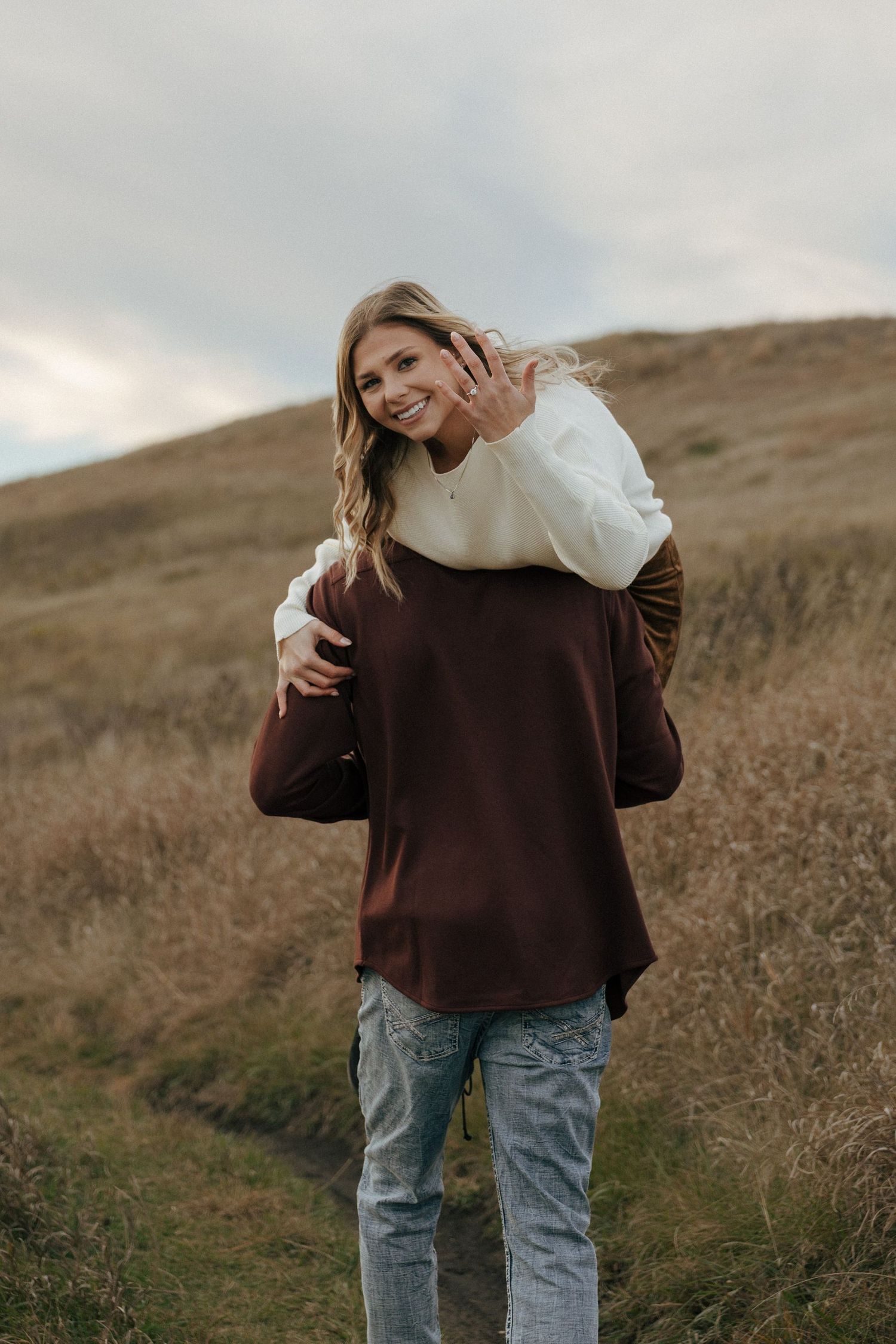 Someone in a brown sweater and jeans poses on a grassy hill slope.