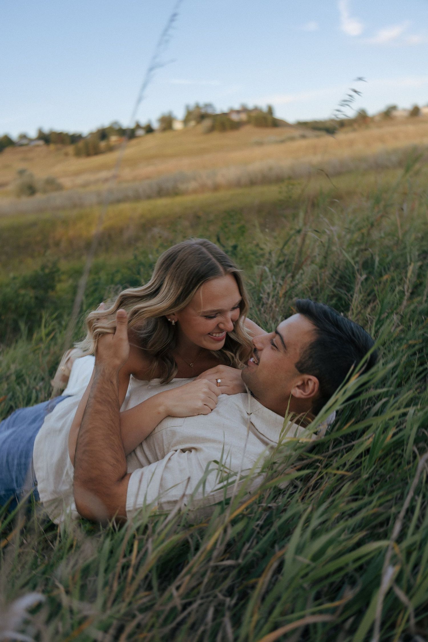 A couple laughs together while lying in a sunlit meadow during a romantic photoshoot.