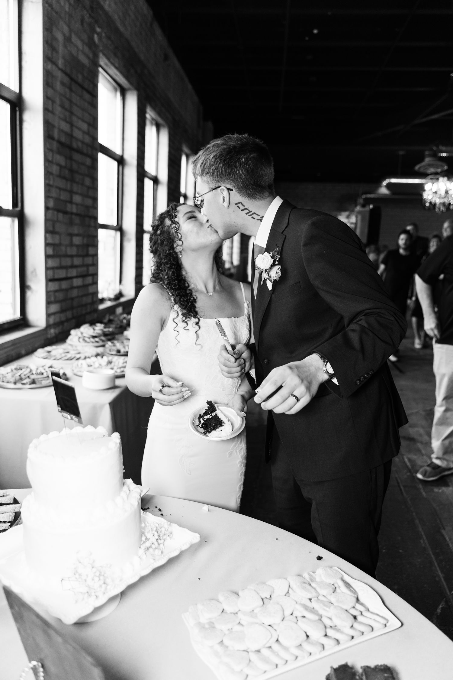Black and white photo of a bride and groom sharing a kiss while cutting their wedding cake at their reception.