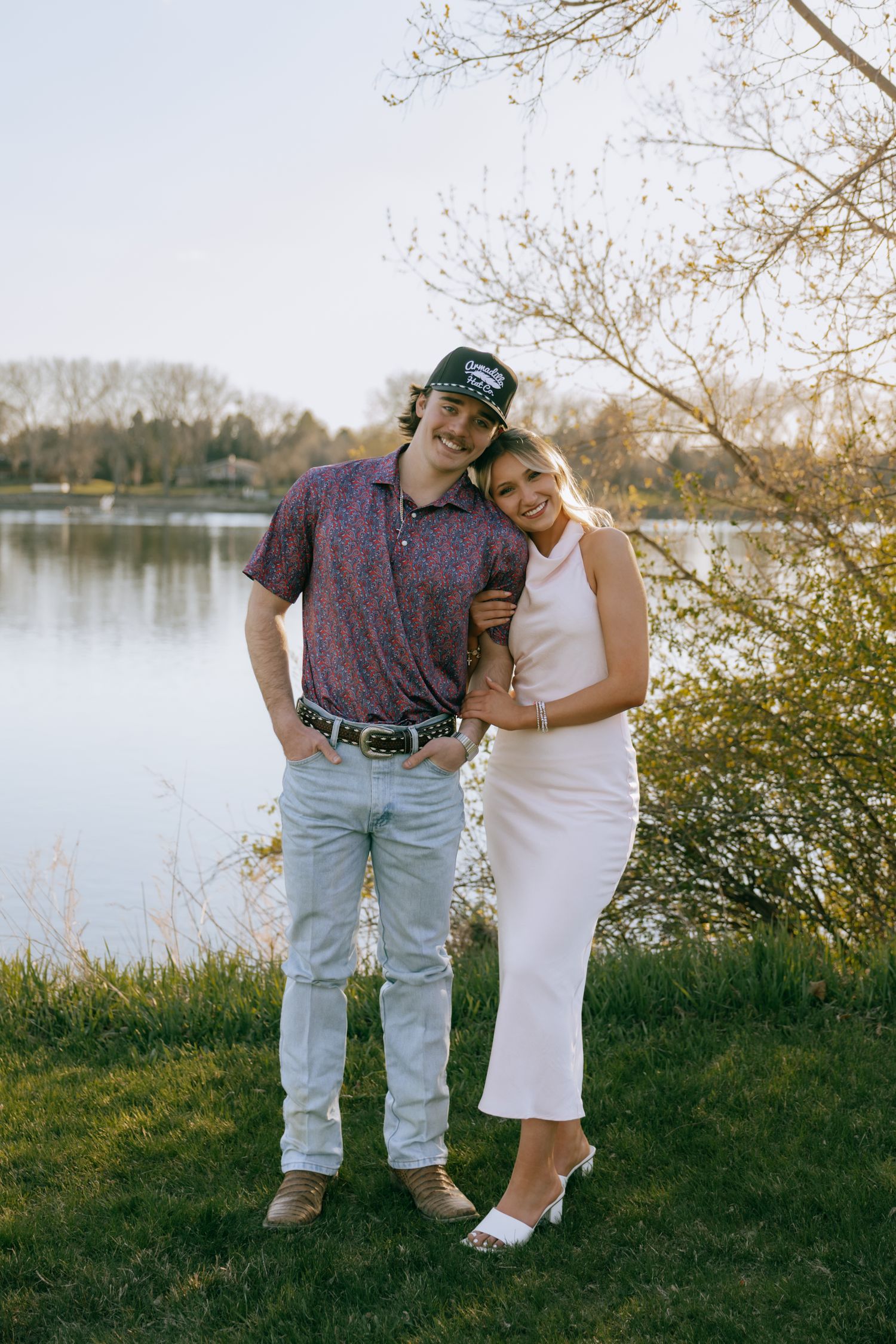 A smiling couple poses by a lakeside in spring, man in polo and cap, woman in white midi dress.