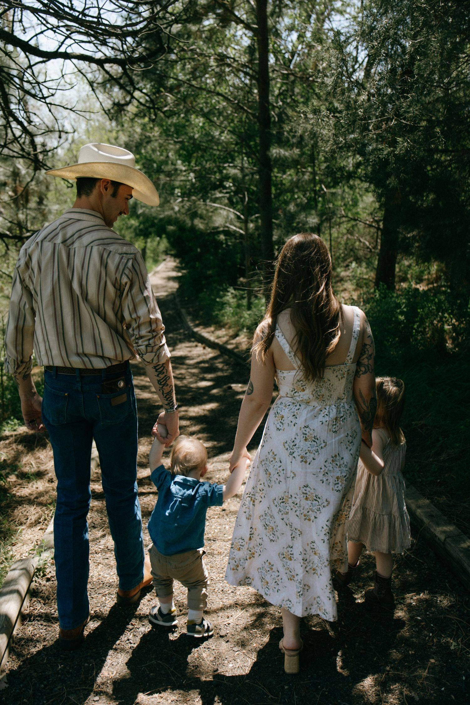 Family of four walking on a forest path, parents holding hands with toddler and young girl in sunny woodland setting.