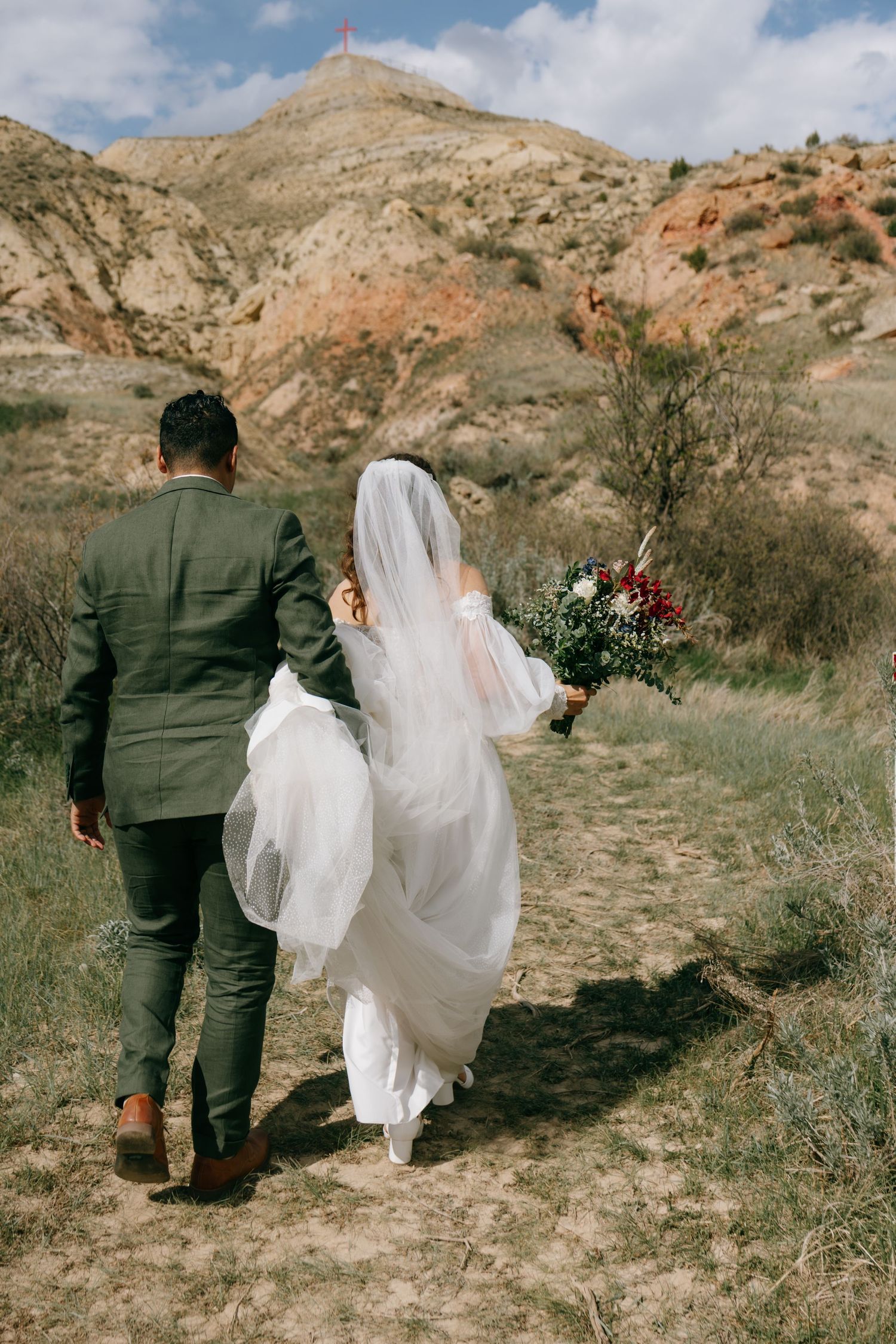 Bride and groom walking hand in hand through a scenic desert landscape, bride holding a bouquet in a flowing white gown.