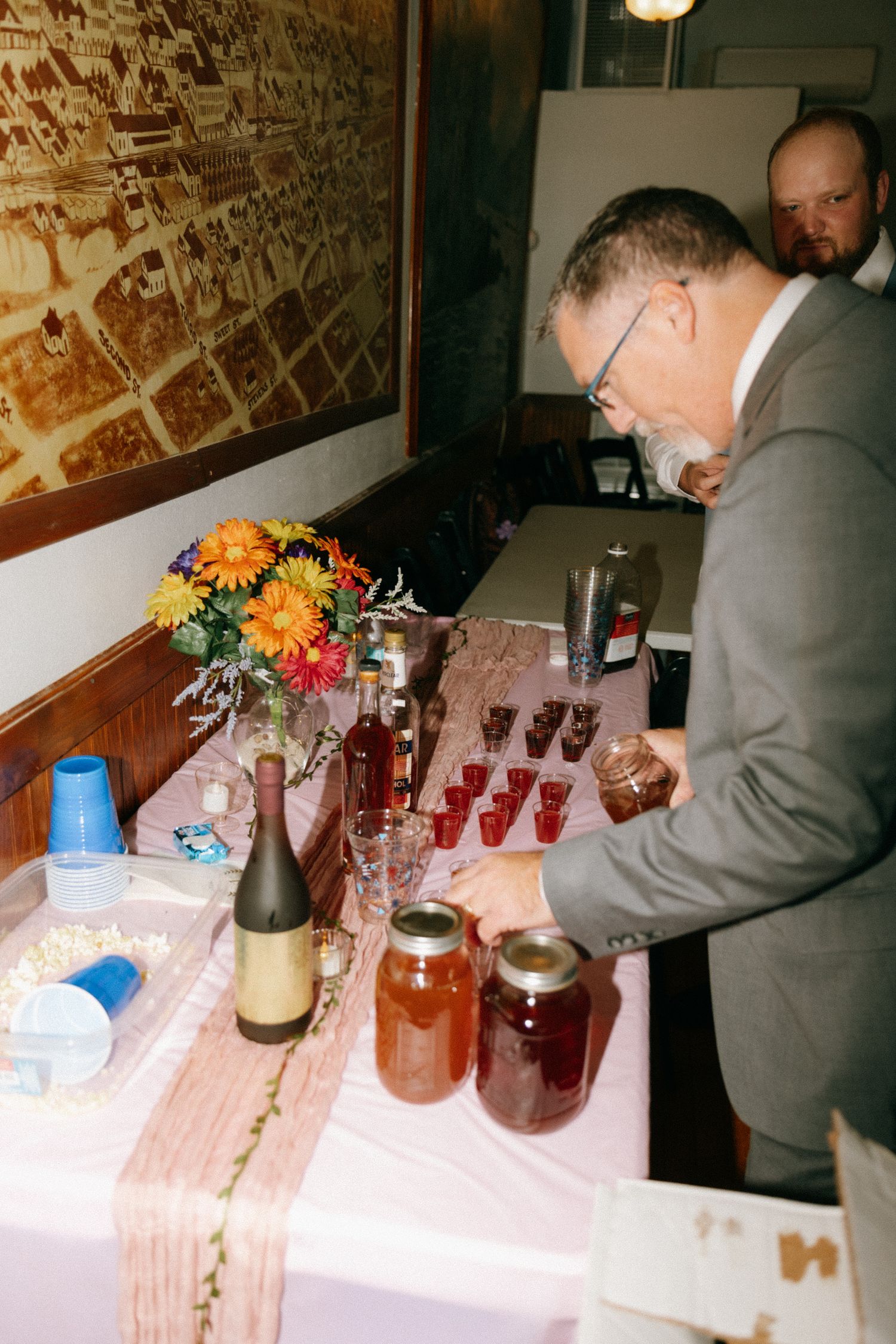 Man in gray suit arranging drinks and mason jars on a decorated table with colorful flowers at an indoor event.