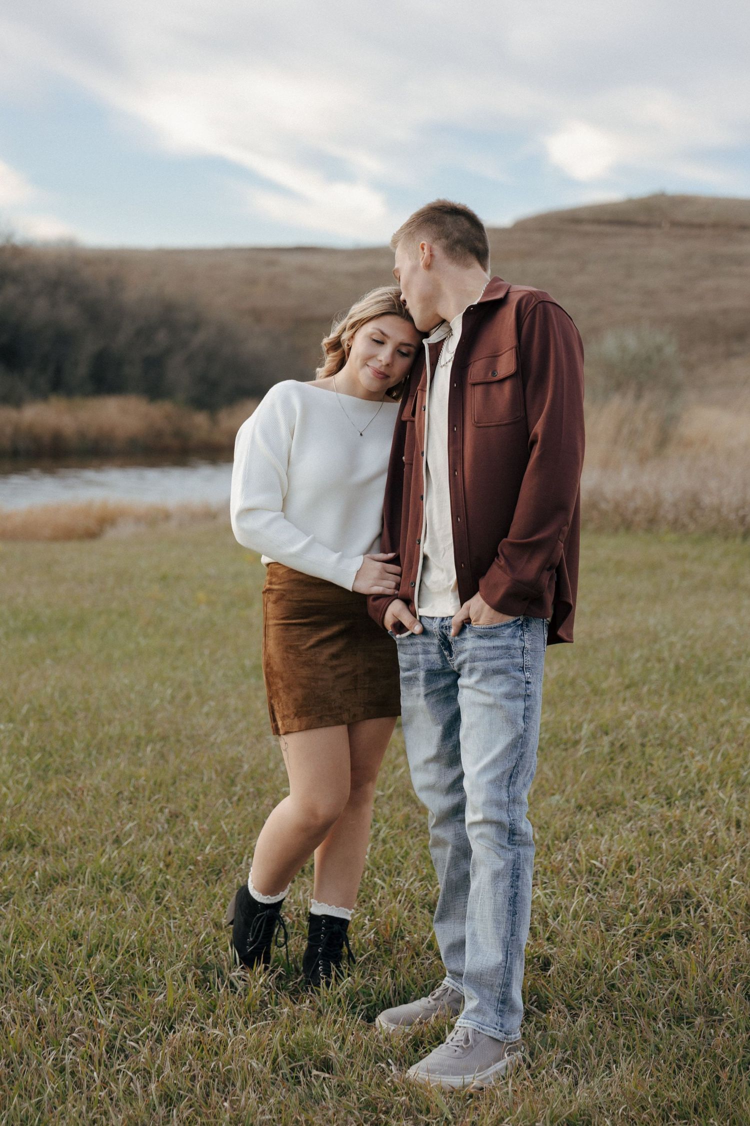 Two people in autumn clothing stand close together in a grassy field with hills in the background.