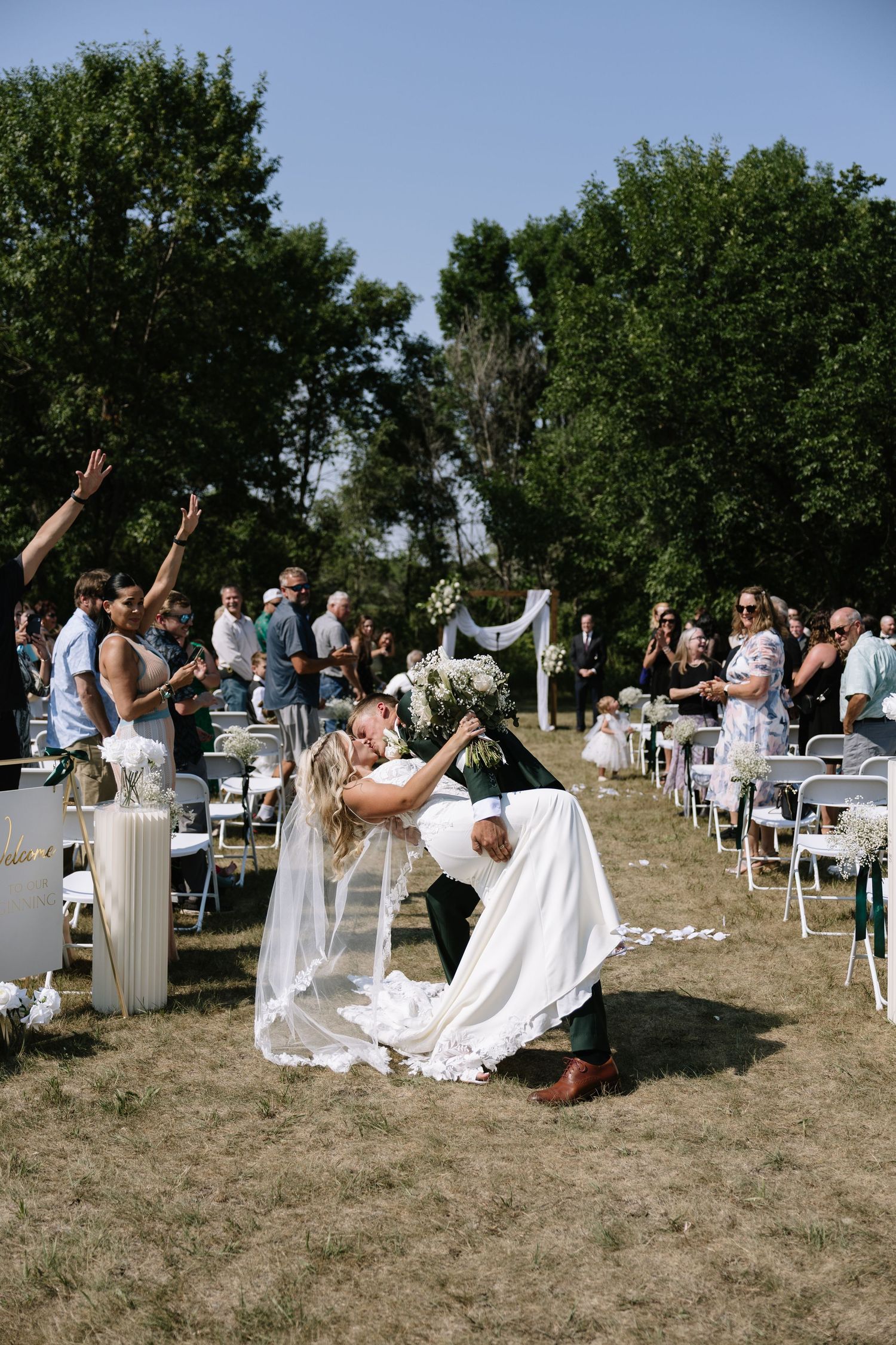 Groom dips bride for a kiss at outdoor wedding ceremony as guests cheer along the aisle.