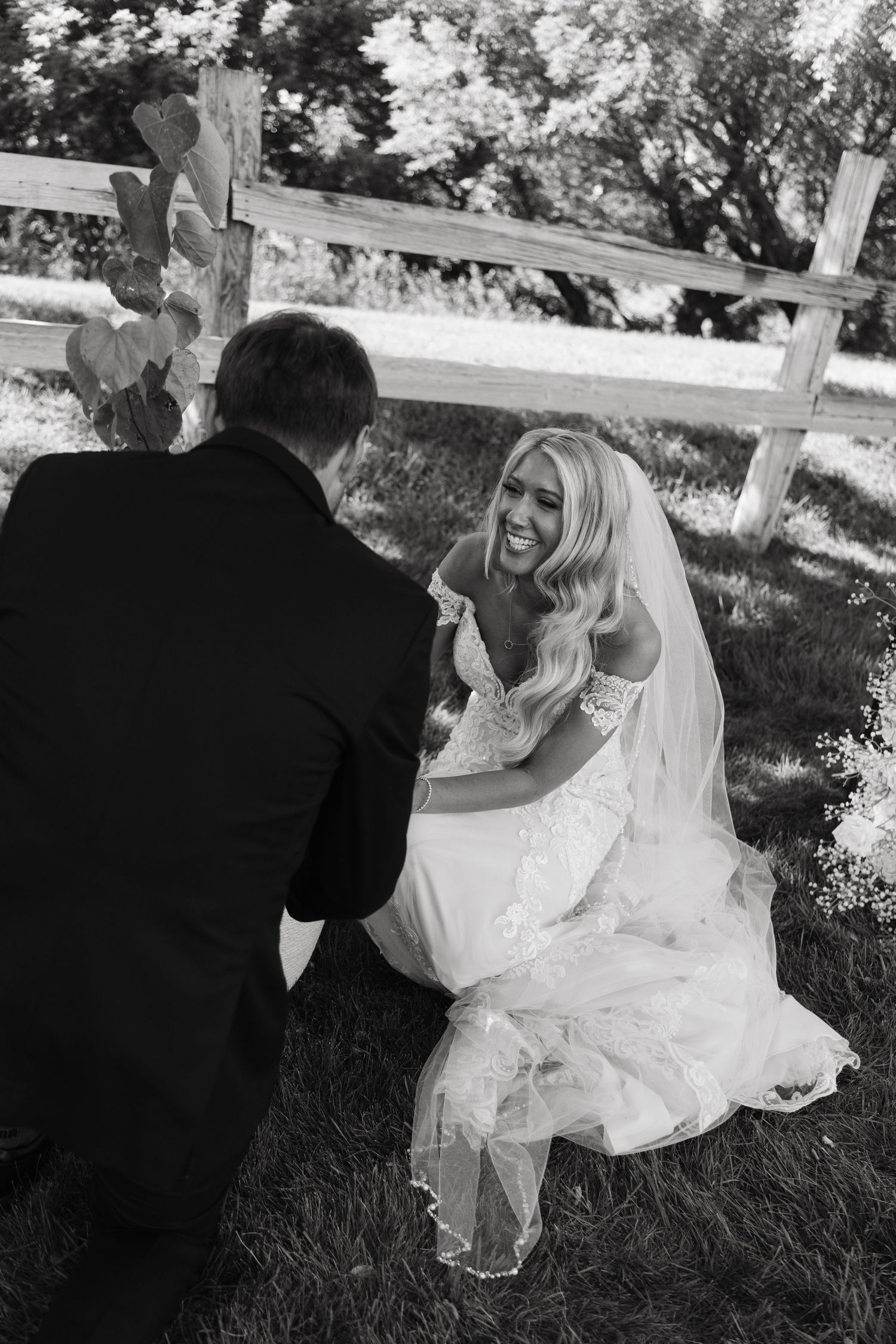 Black and white photo of a smiling bride in a lace gown and veil kneeling with her groom outdoors near a wooden fence.