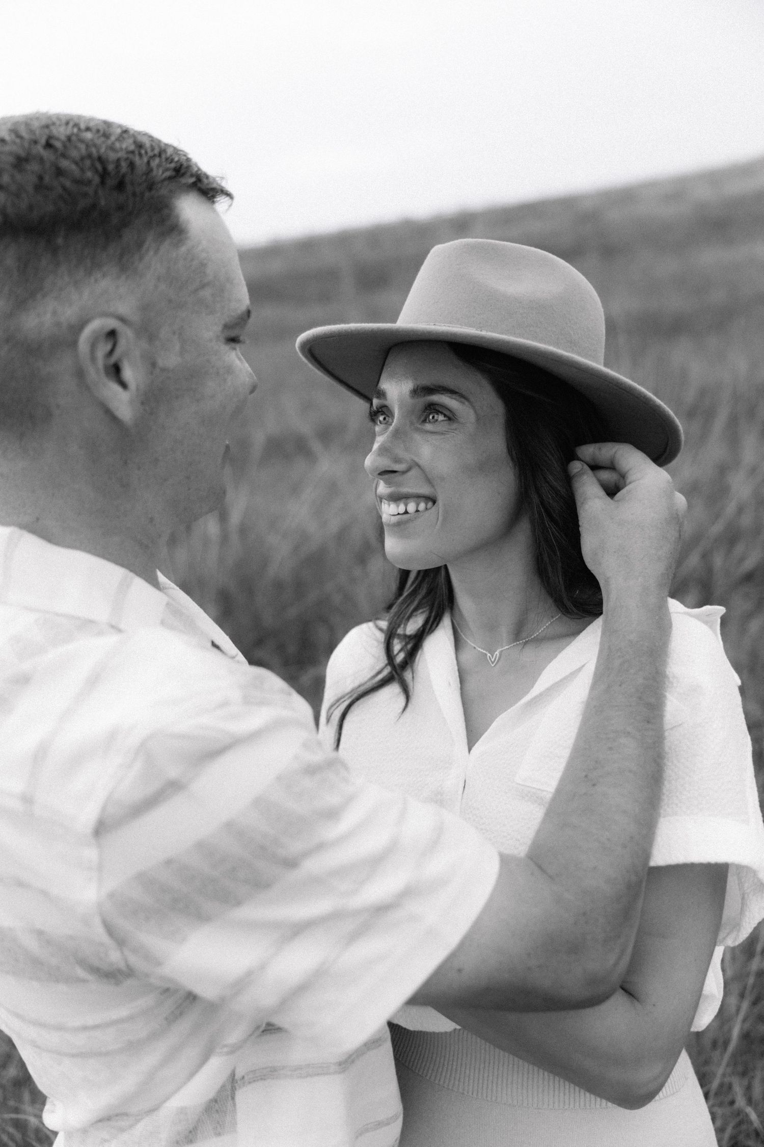 Black and white photo of a smiling woman in a hat being embraced by a man in a field.