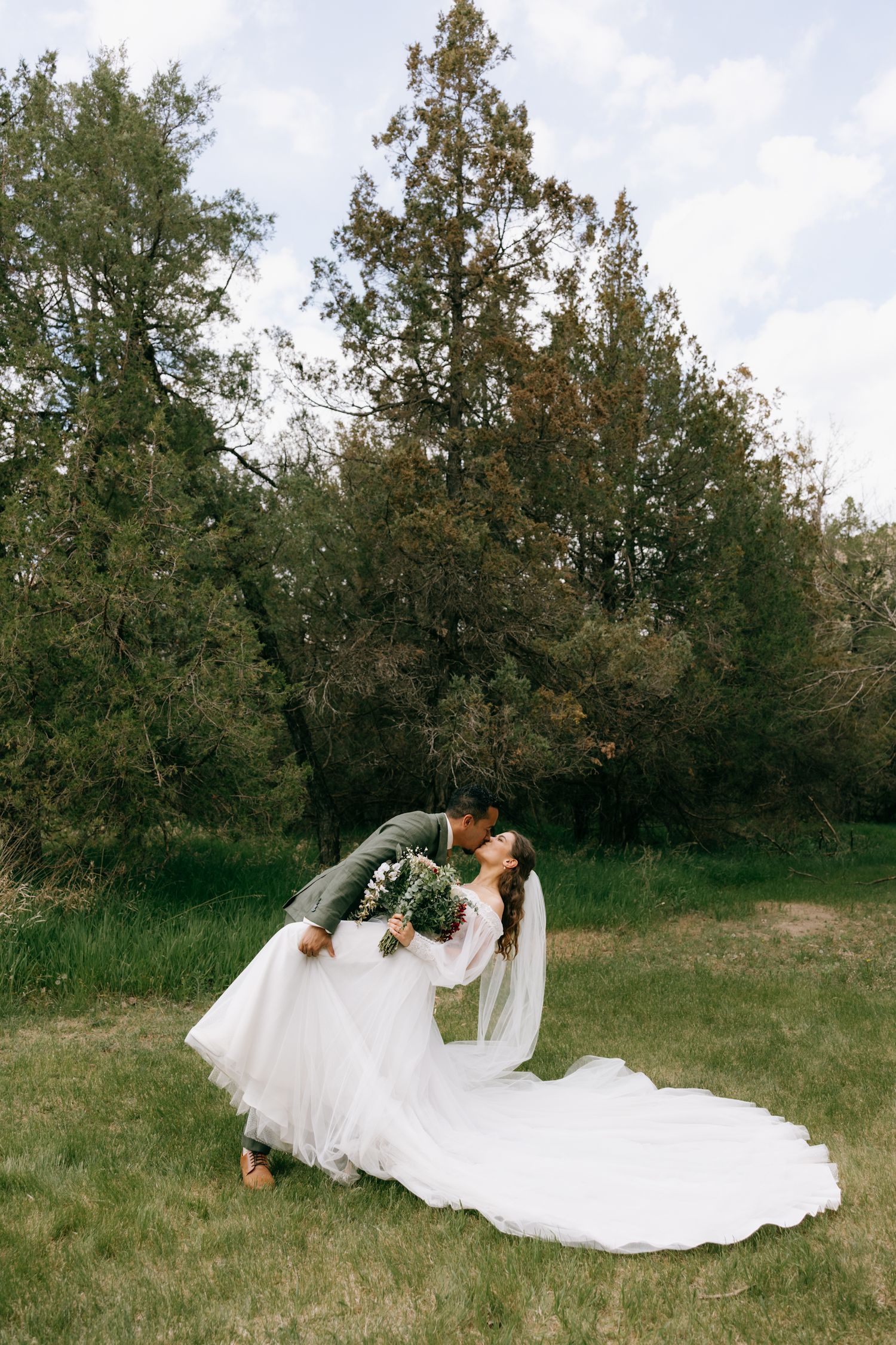 Groom dips bride for a romantic kiss outdoors, her white gown flowing on green grass with tall trees behind them.