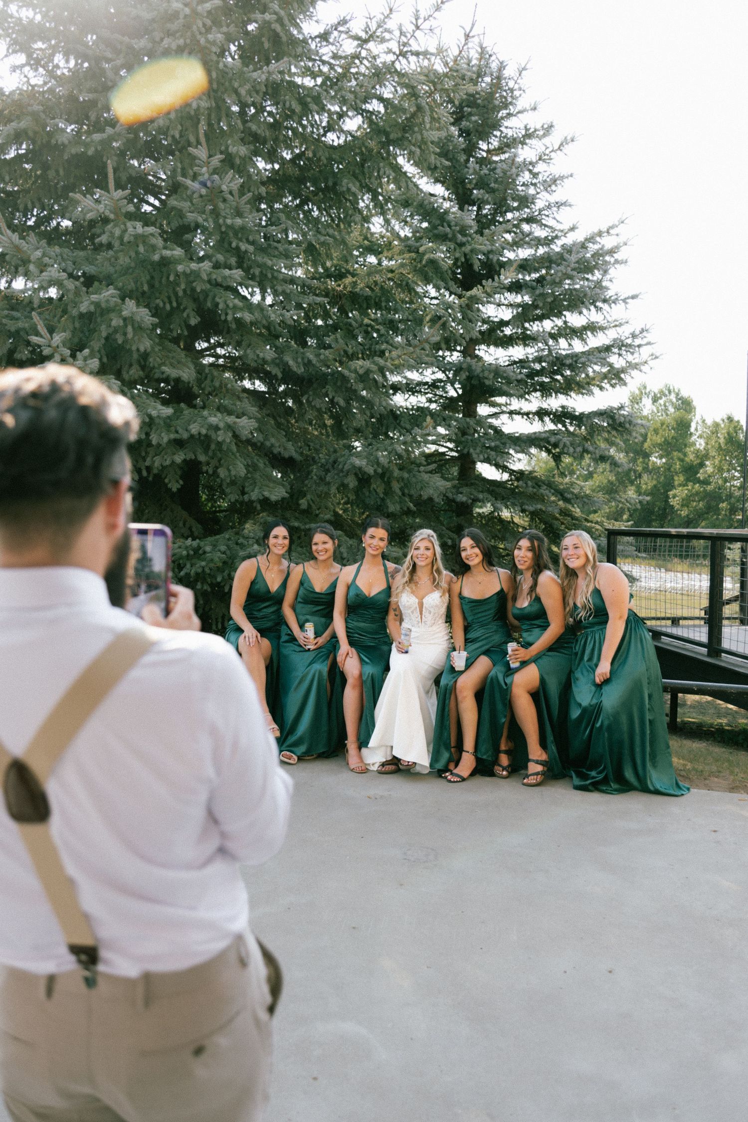 A groom photographs his bride and bridesmaids in emerald green gowns posing outdoors near a large evergreen tree.