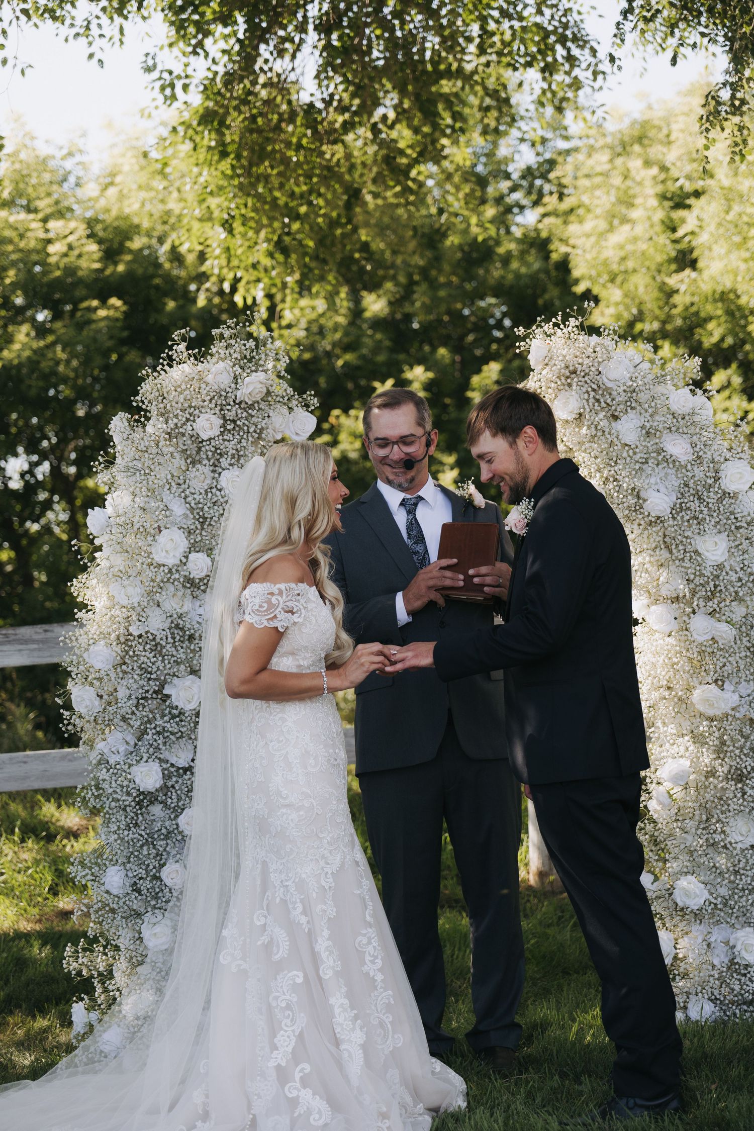 Bride and groom exchanging rings at outdoor wedding ceremony under floral arch with baby's breath and white roses.