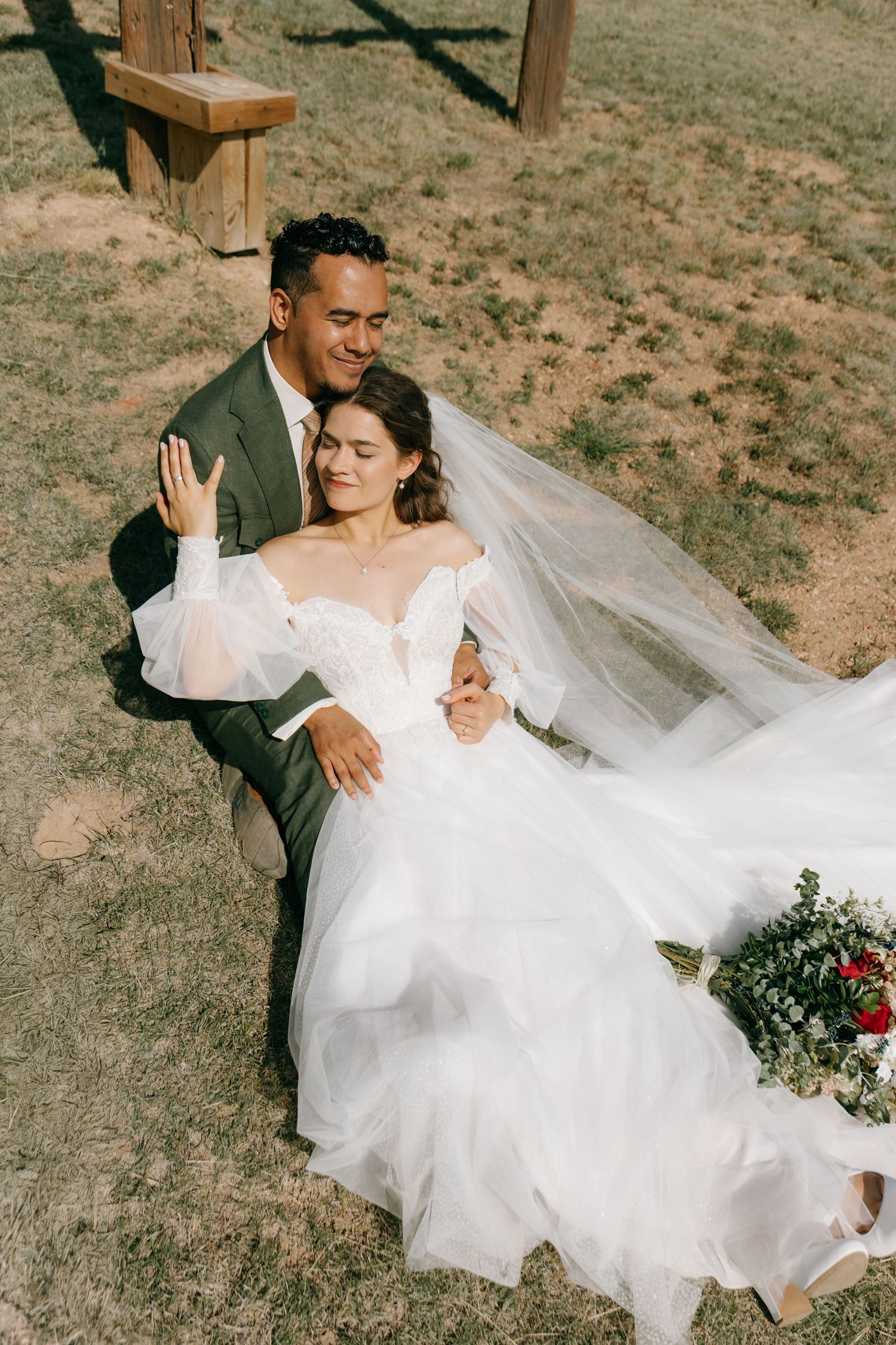 Bride in white off-shoulder gown and groom in dark suit sit together outdoors, sharing a tender moment on their wedding day.