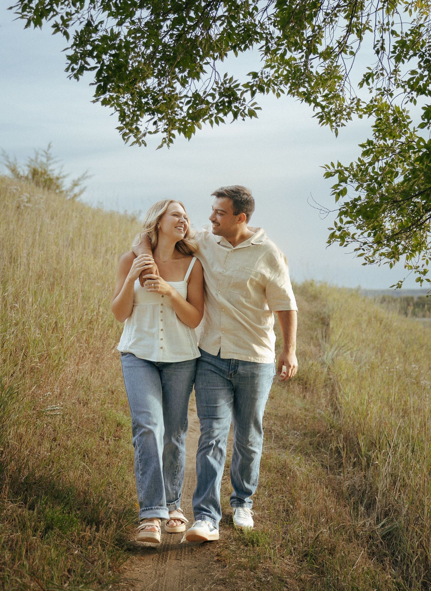 A couple walking together on a grassy hillside path wearing jeans and white shirts on a sunny day.