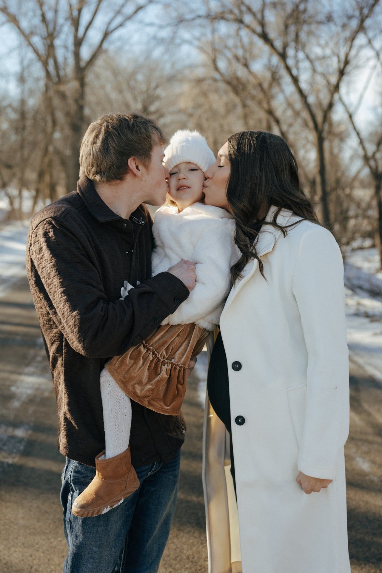 photos of mom and dad kissing cheeks of daughter in snowy trees in Bismarck