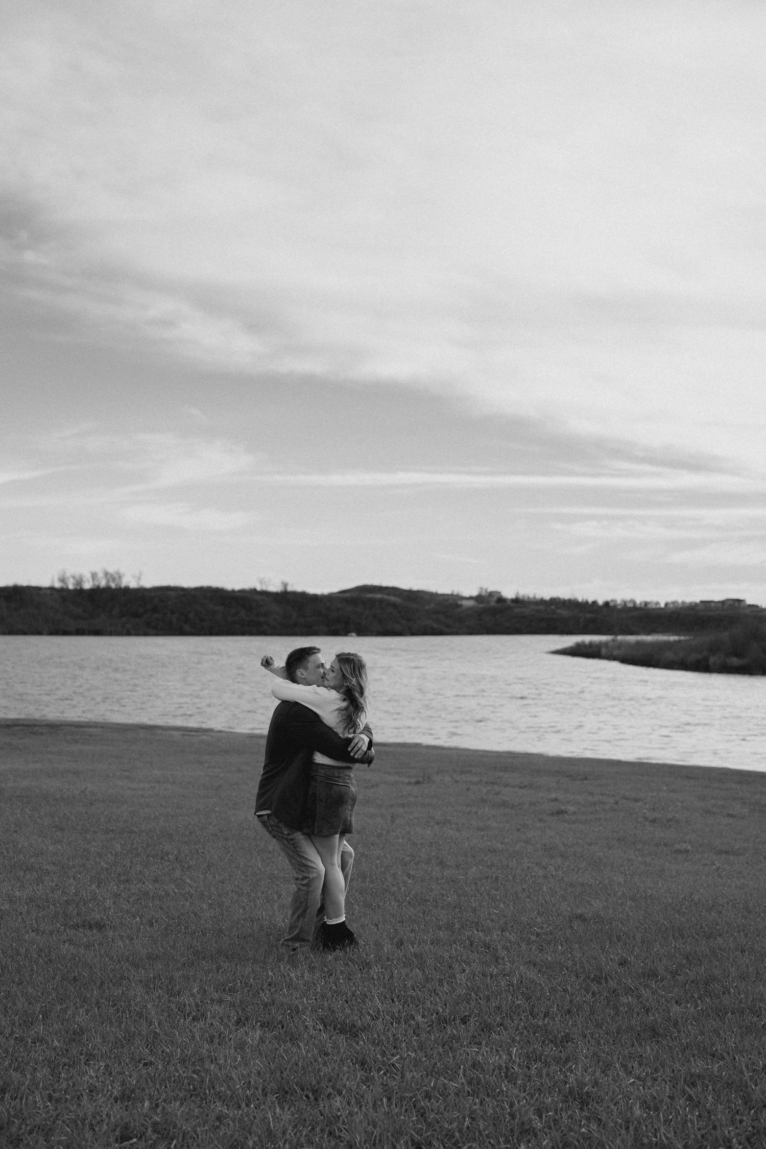 A black and white photo captures a couple embracing in a field with water and hills in the distance.