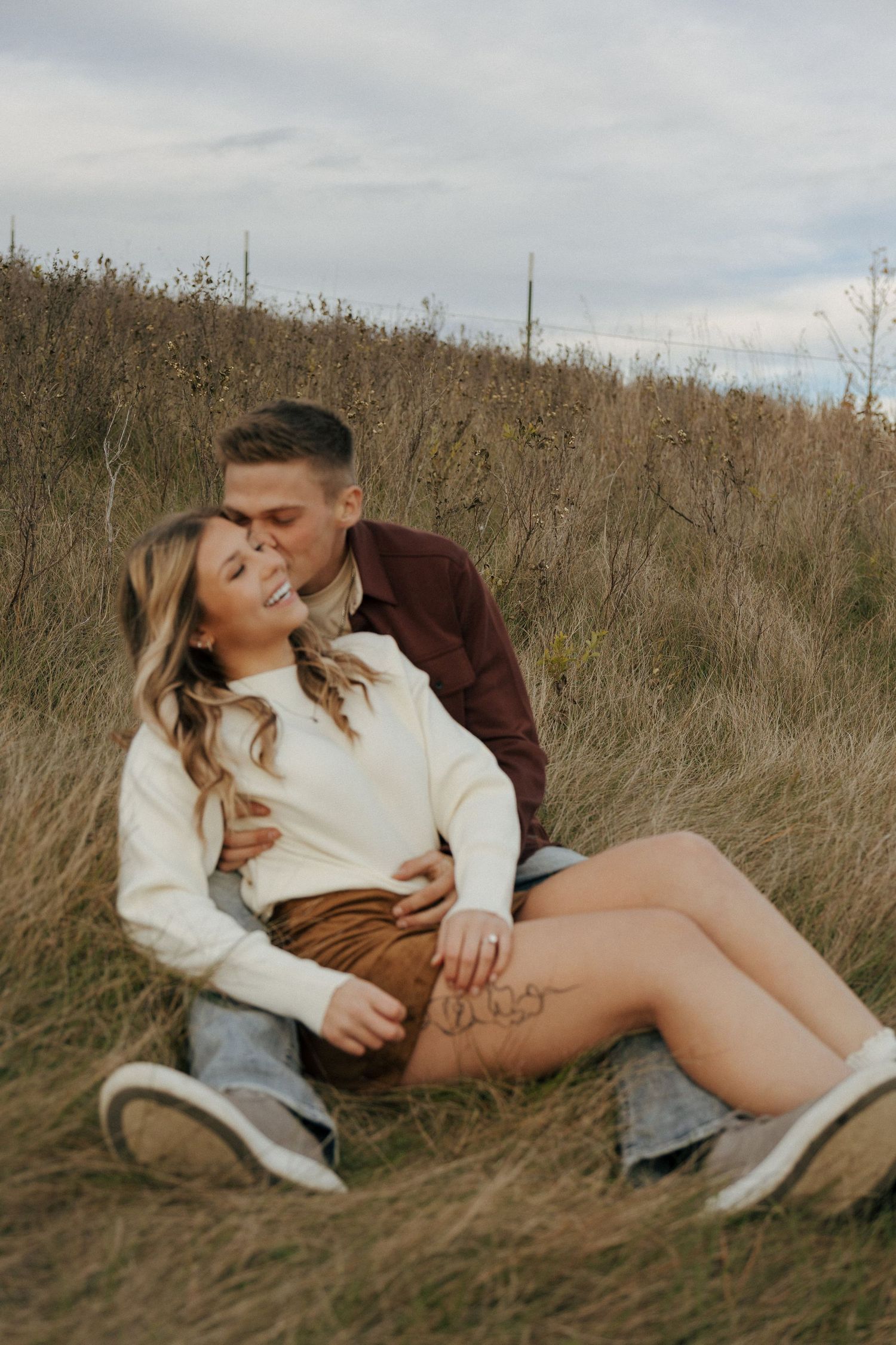 Couple lounging in dry grass field under cloudy sky.