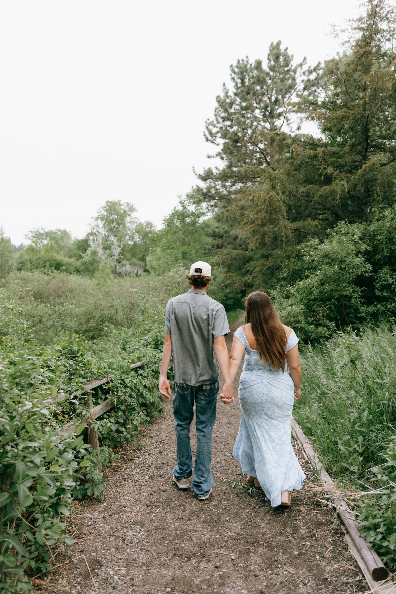 A couple walks hand in hand along a nature trail surrounded by lush greenery, viewed from behind.