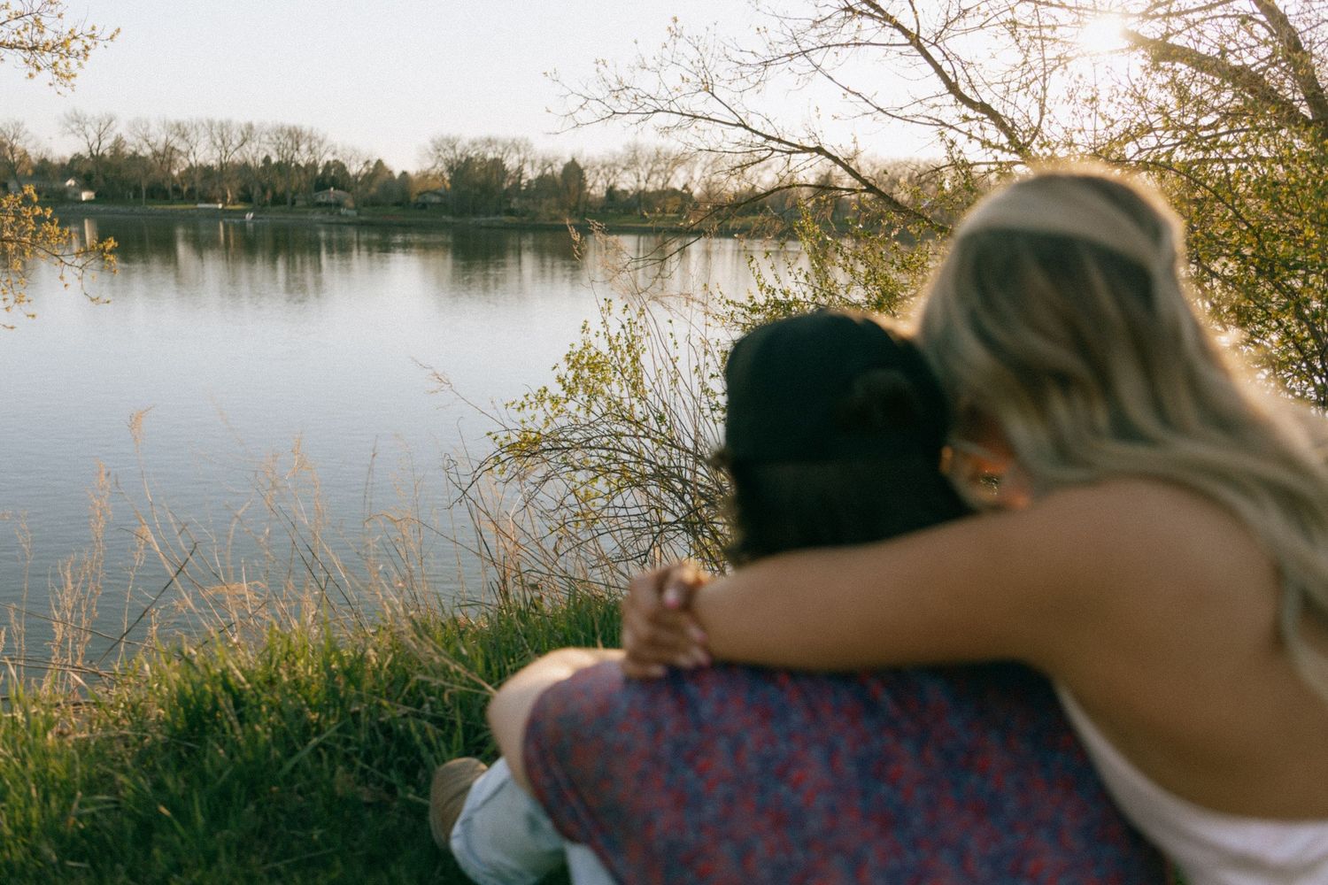 Two people sitting together by a peaceful lake, embracing while enjoying the scenic waterfront view surrounded by trees.