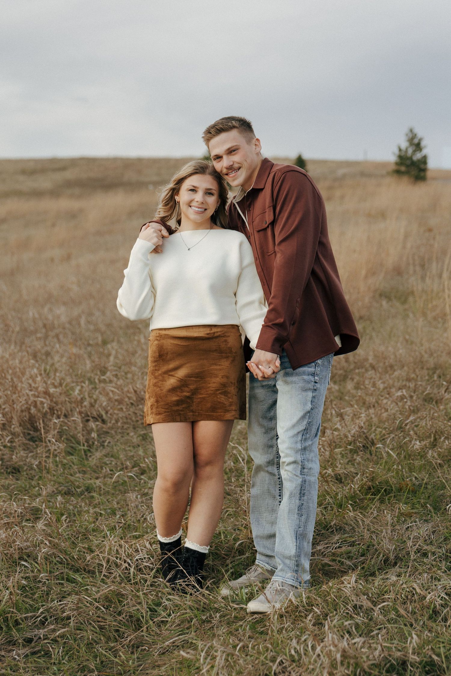 Couple poses together in casual autumn outfits against rolling hills landscape.